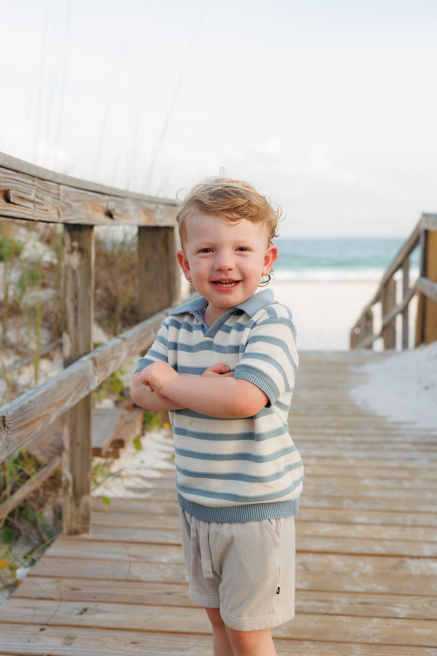 A smiling toddler boy in a striped shirt stands on a wooden beach boardwalk with ocean waves in the background.