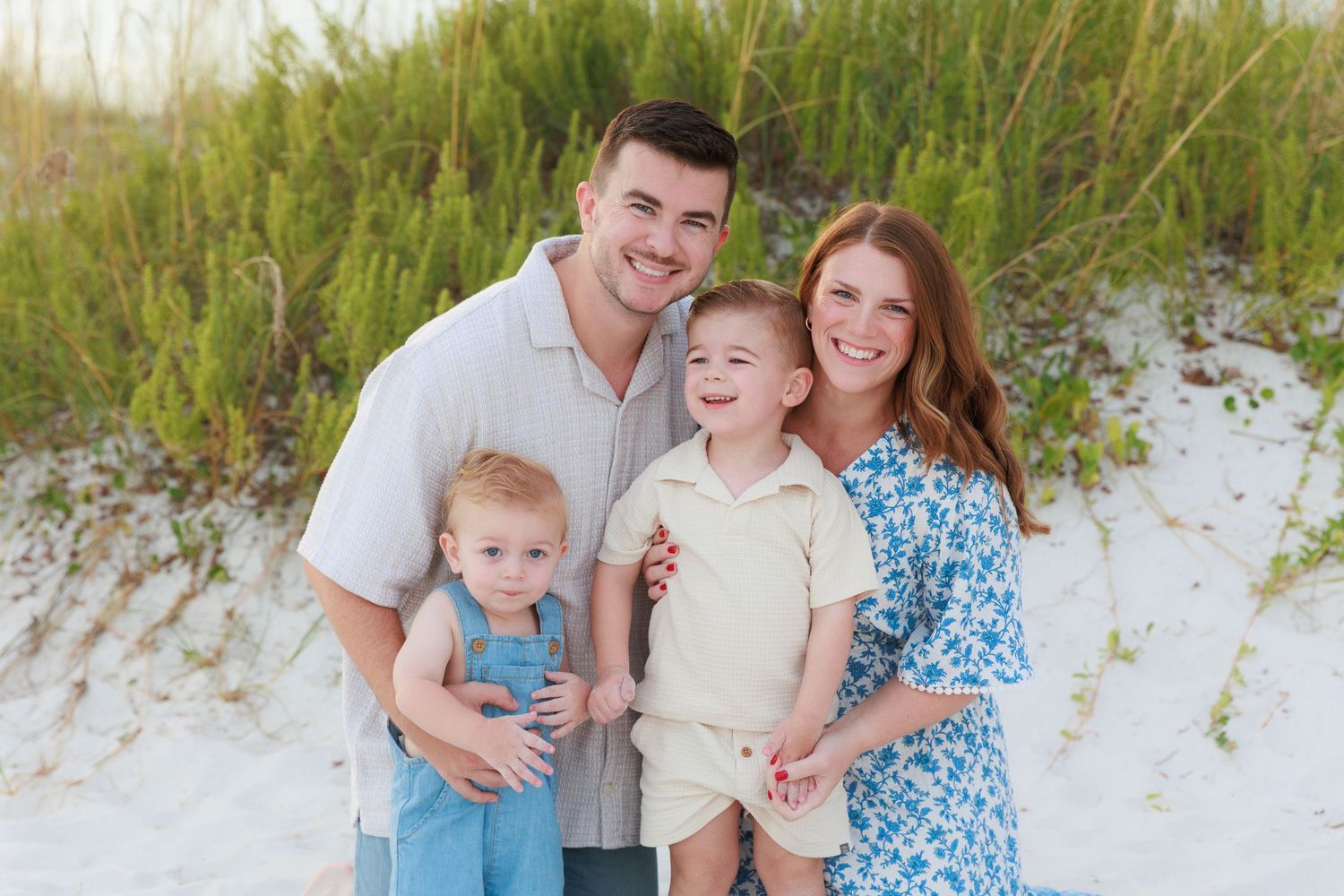 Family of four smiling together on a sandy beach with green dunes, parents with two young boys in summer outfits.