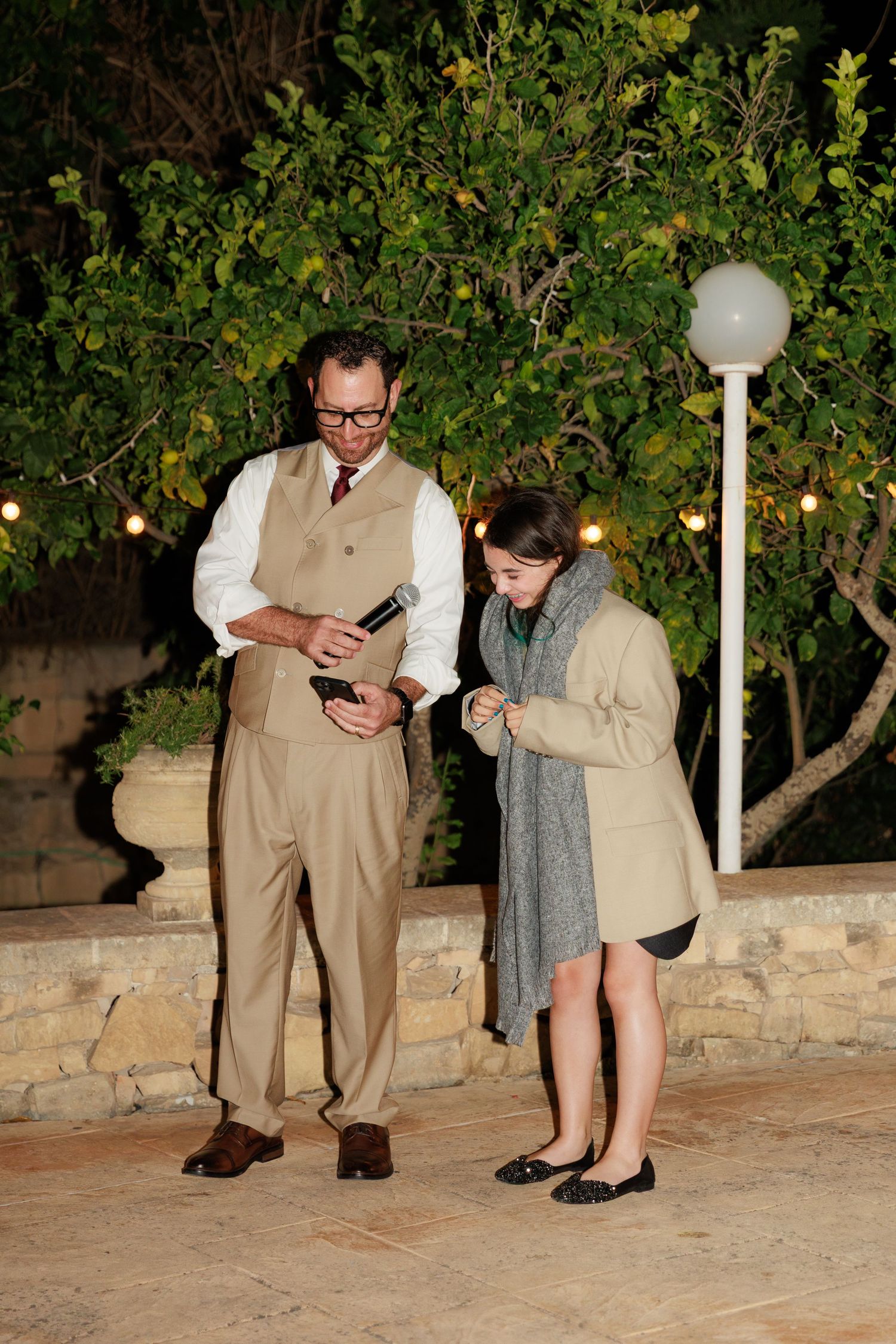 Two people looking down at their phones while standing outdoors at night near decorative lighting and greenery.