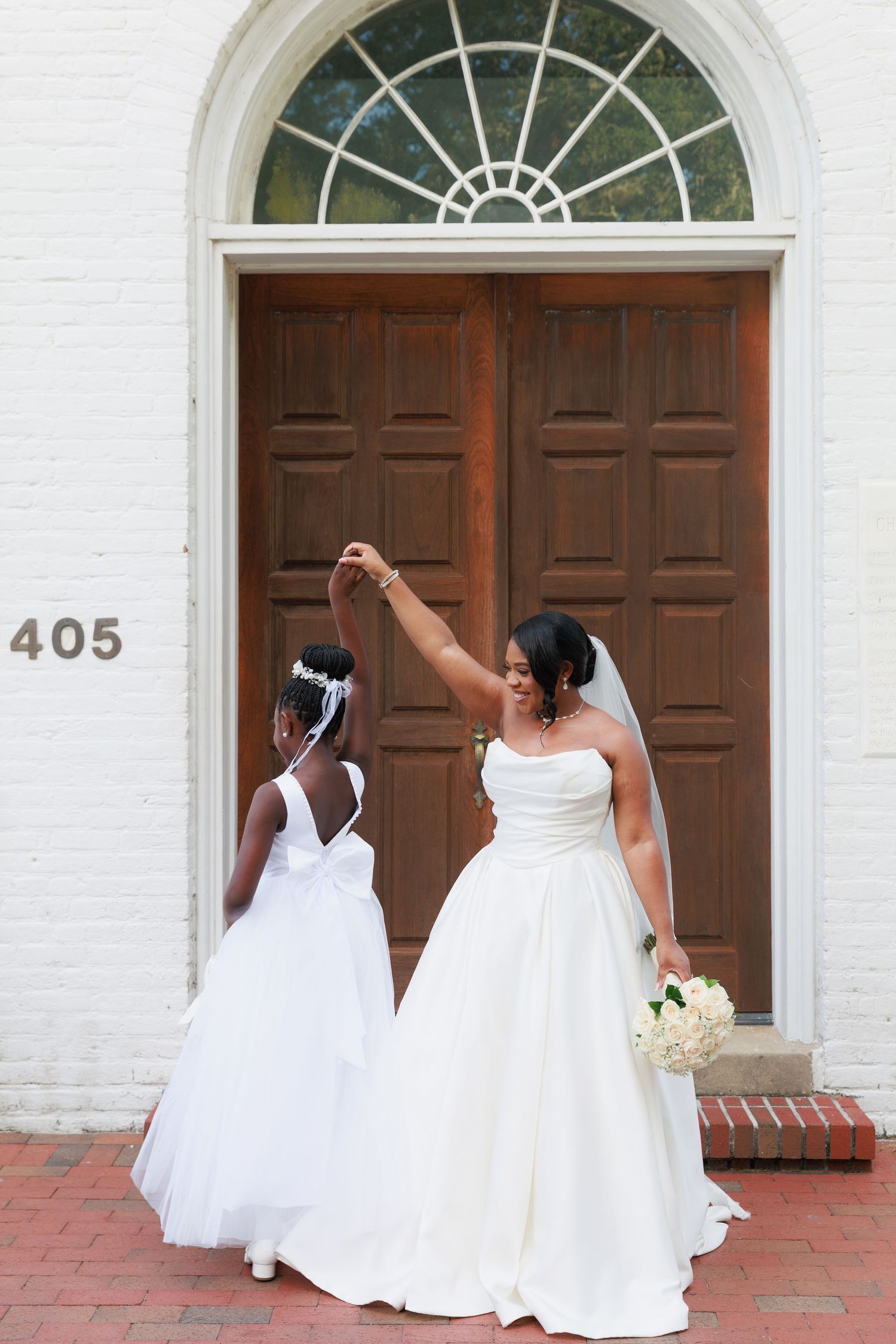 Two brides in elegant white wedding gowns pose playfully in front of a historic brick building with wooden doors.