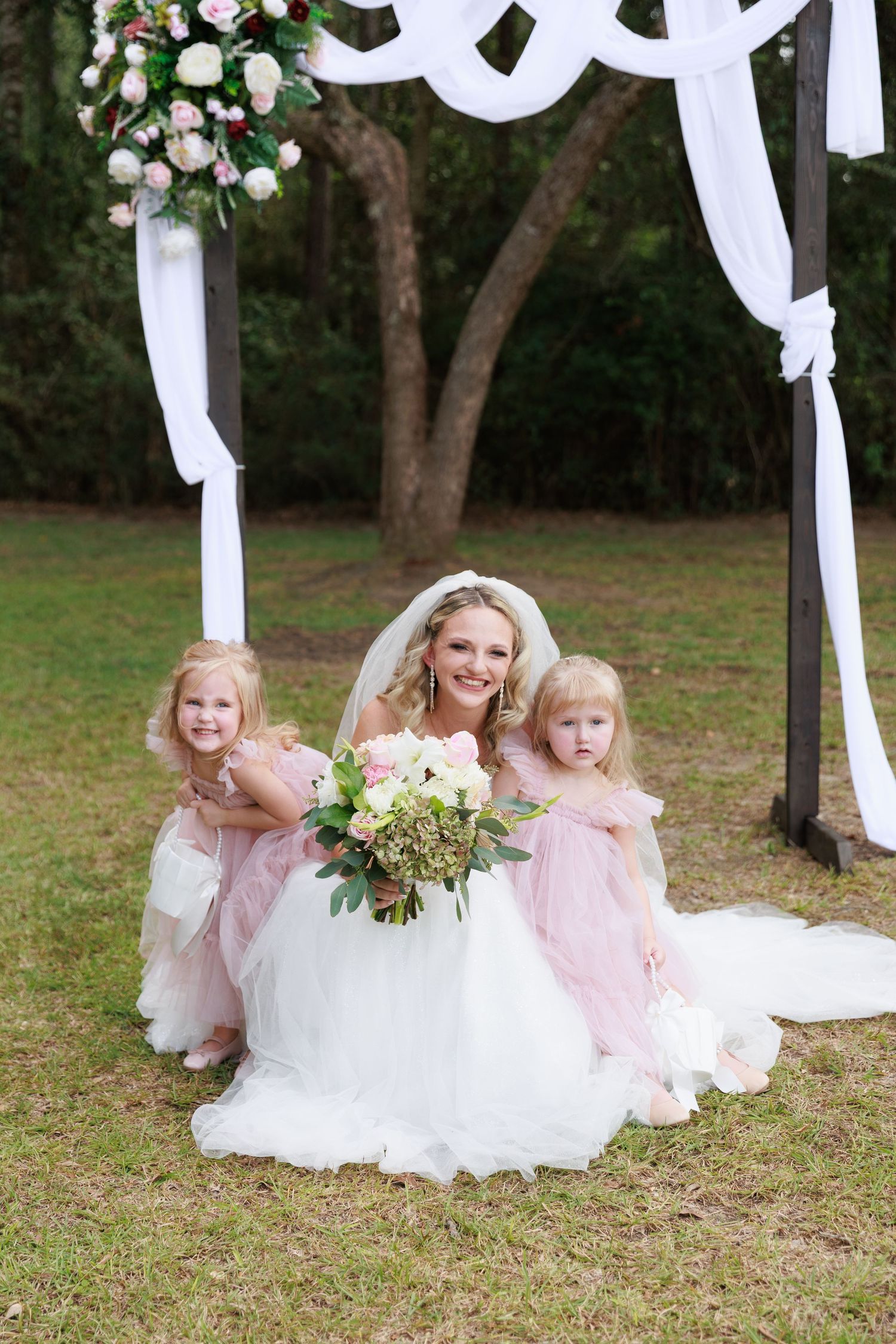 Wedding party members in pink and white dresses sit gracefully on grass beneath a white draped arch with floral accents.