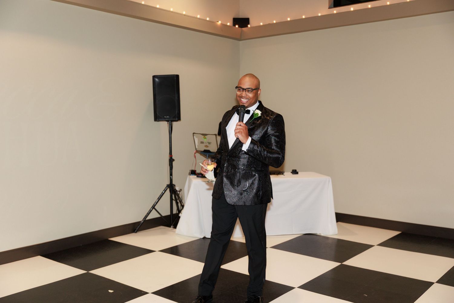 A speaker in a black suit holds a microphone while standing on a black and white checkered dance floor.