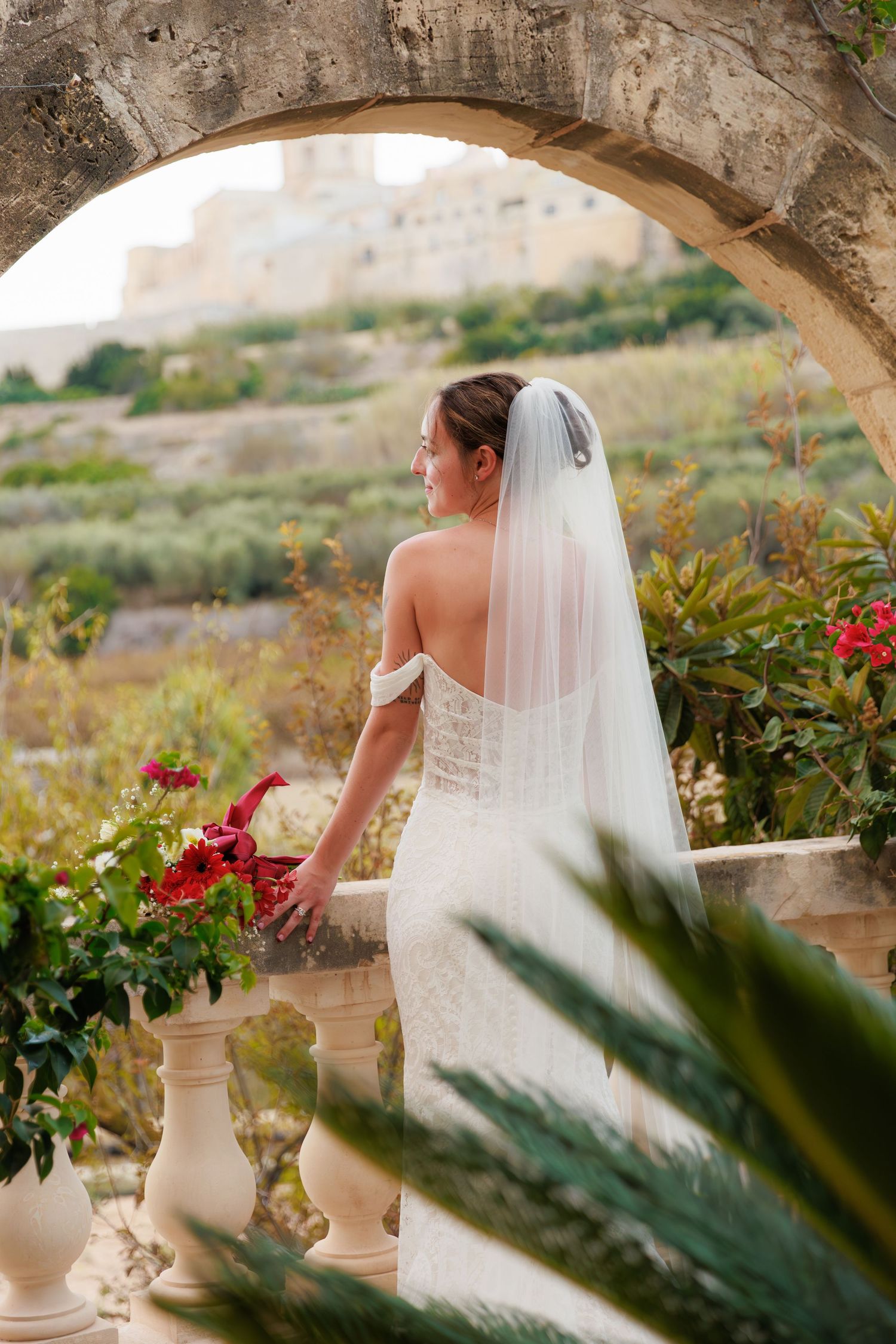 A bride in a white dress and veil stands by stone archway with colorful flowers.
