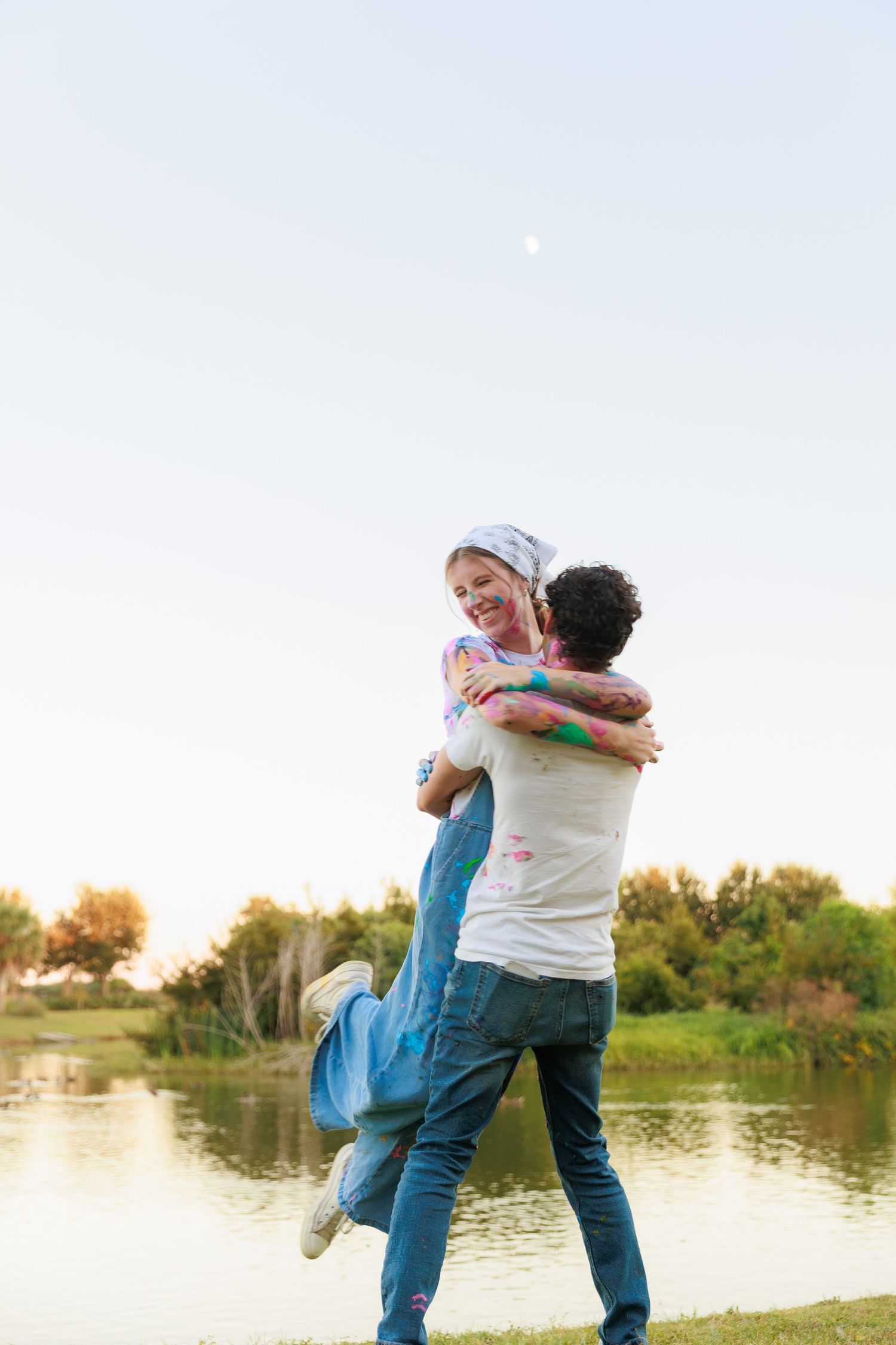 Couple in casual attire playfully embrace and spin by a serene lake during golden hour.