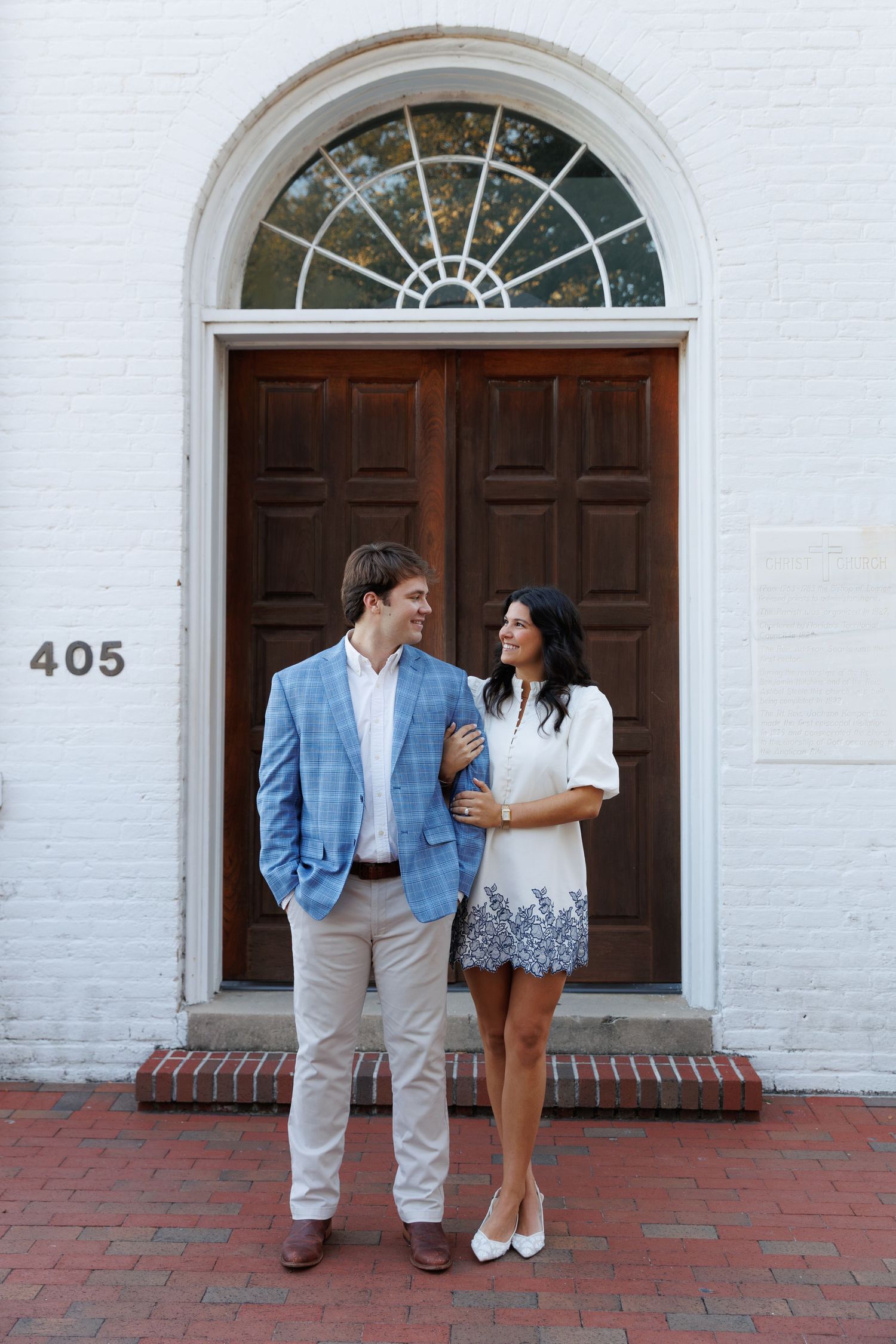 A couple stands together in front of white brick walls and arched wooden doors on a red brick sidewalk.