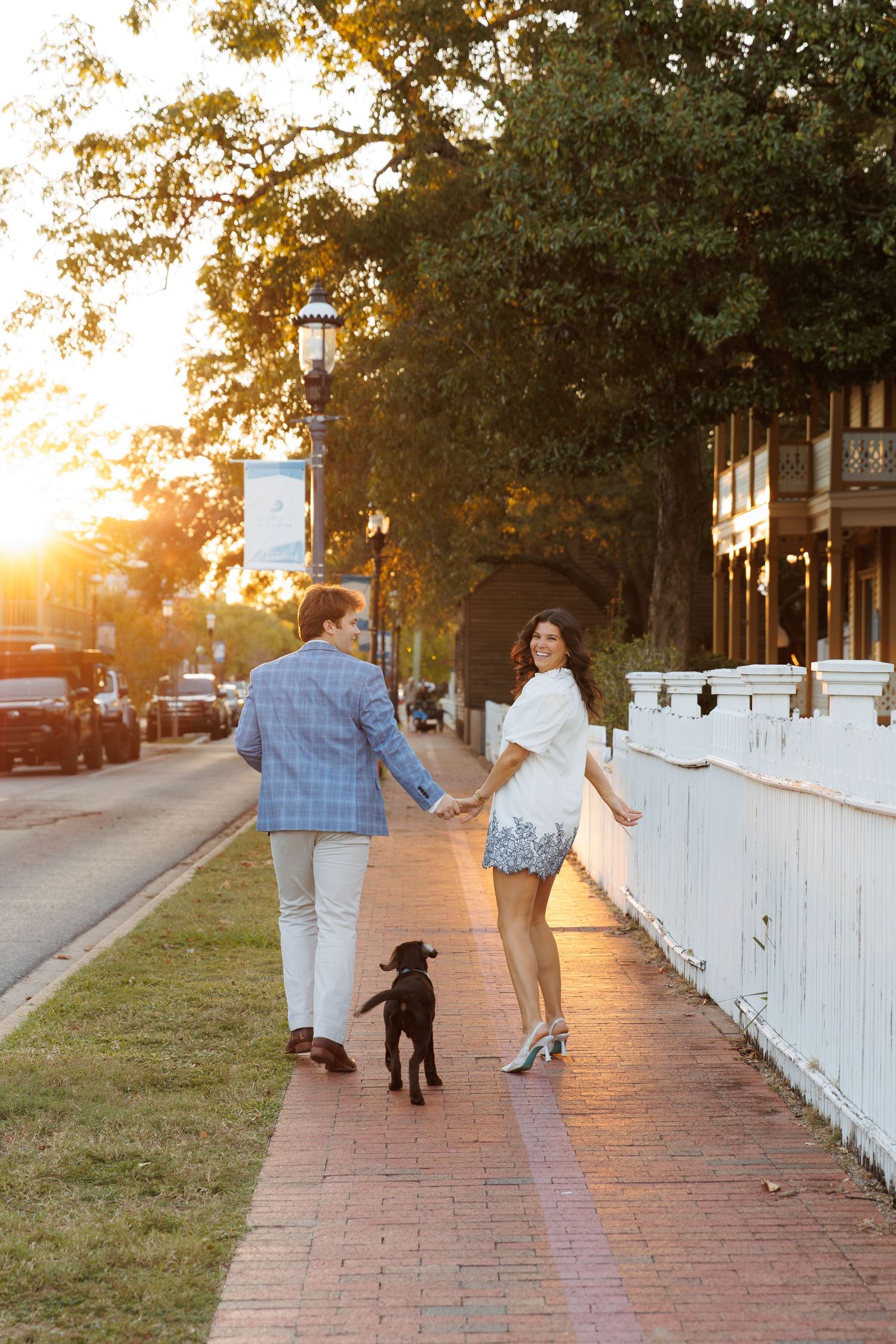Family walking along a brick sidewalk with white picket fence at sunset creating warm golden lighting.