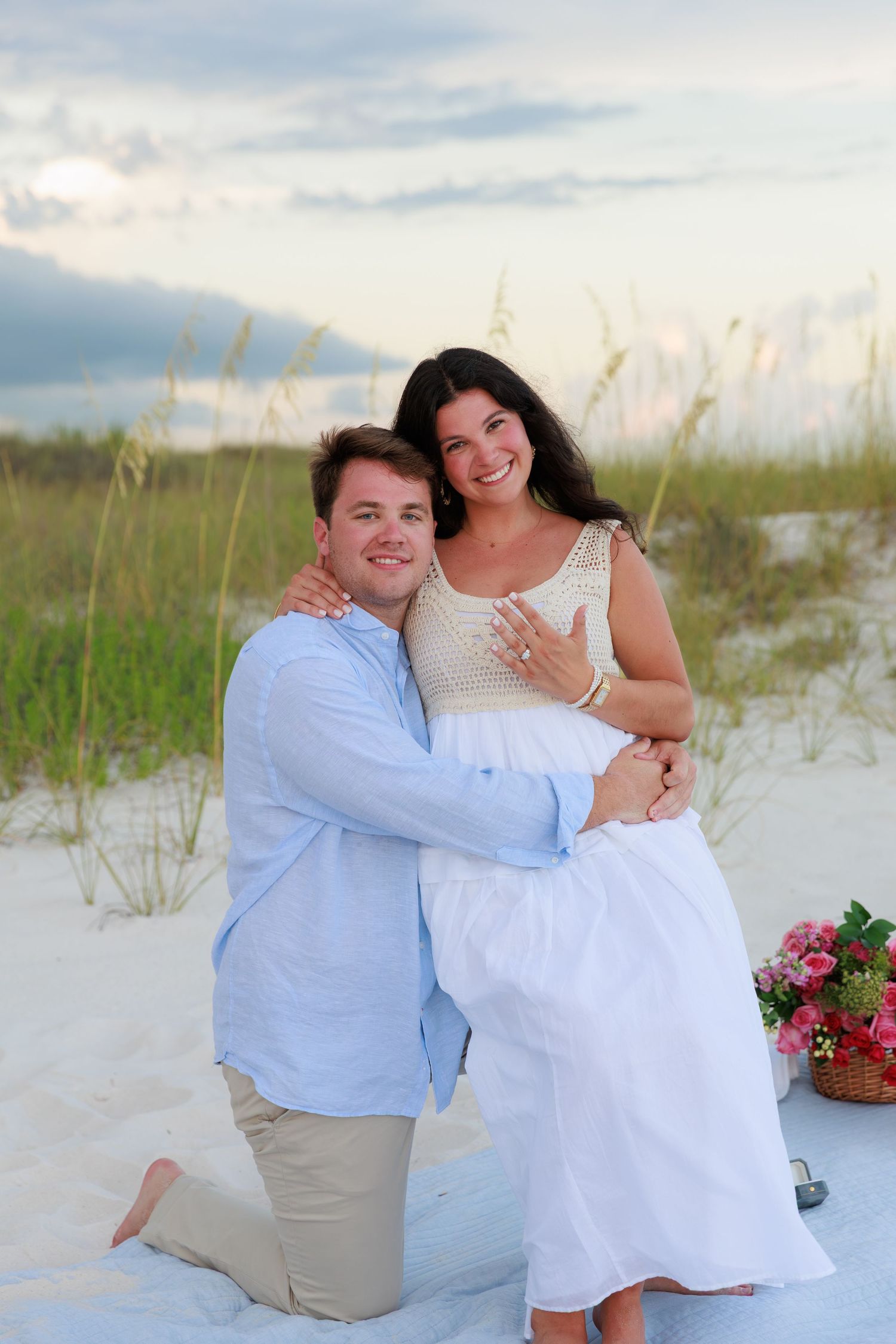 A couple shares a romantic moment on a white sandy beach with sea oats and pink flowers at sunset.