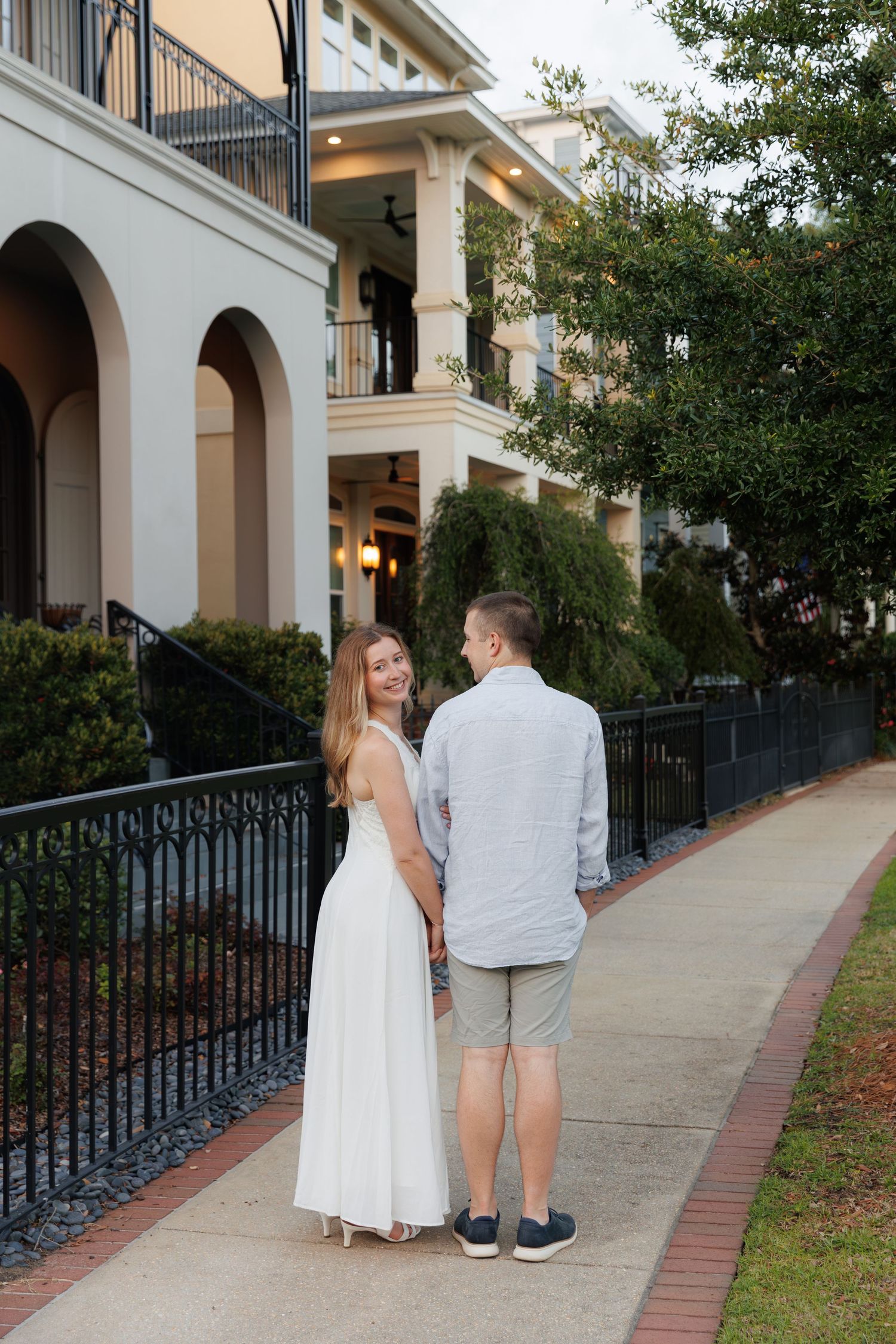 A couple holds hands on a sidewalk, woman in white dress smiling back, elegant building with arches behind them.