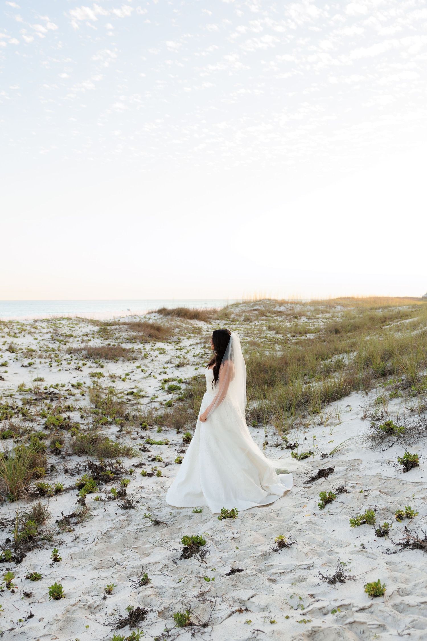 Bride in white gown and veil standing on sandy beach dunes with sea grass and ocean in background.