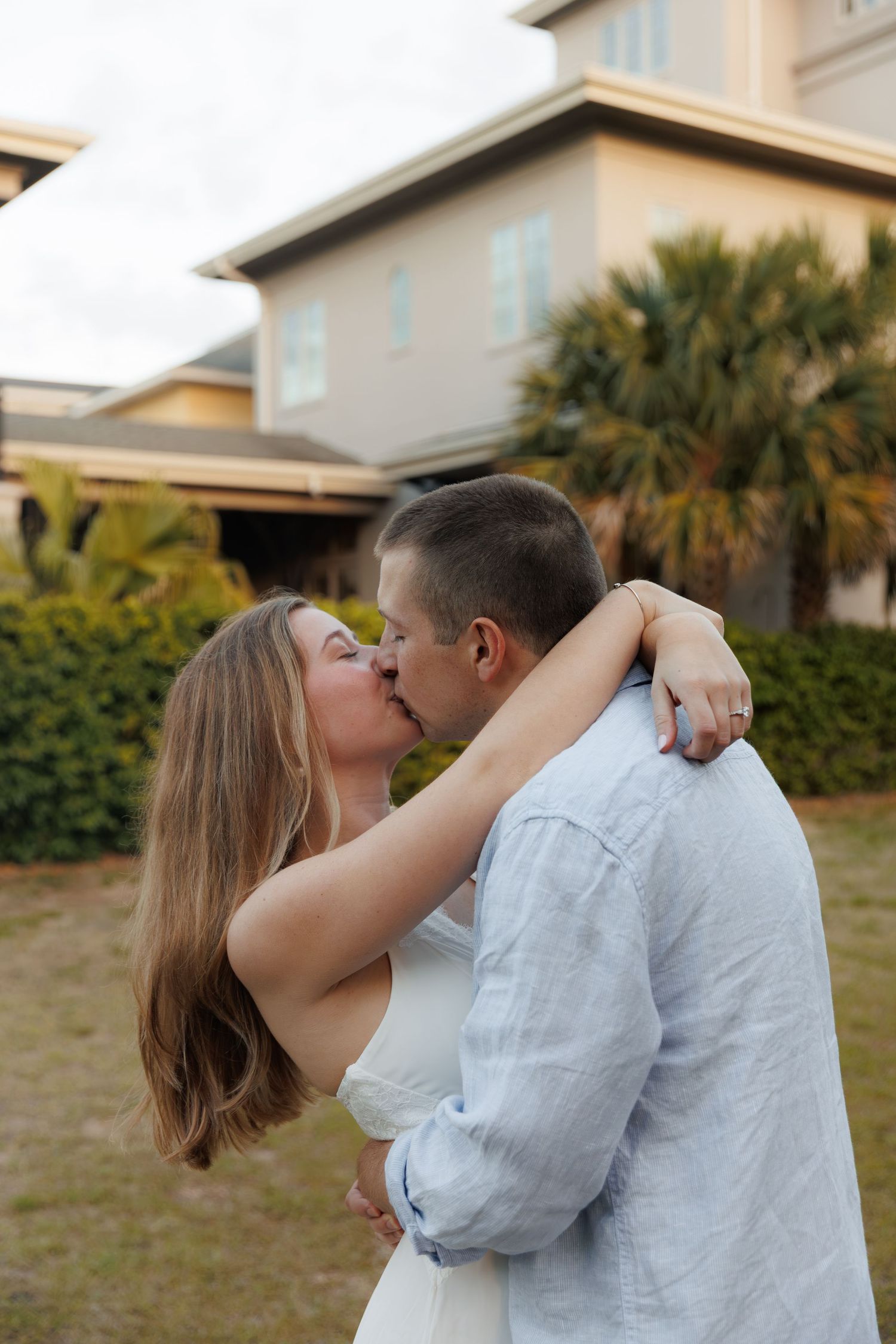 Couple sharing a romantic kiss outdoors with a large coastal home and palm trees in the background.