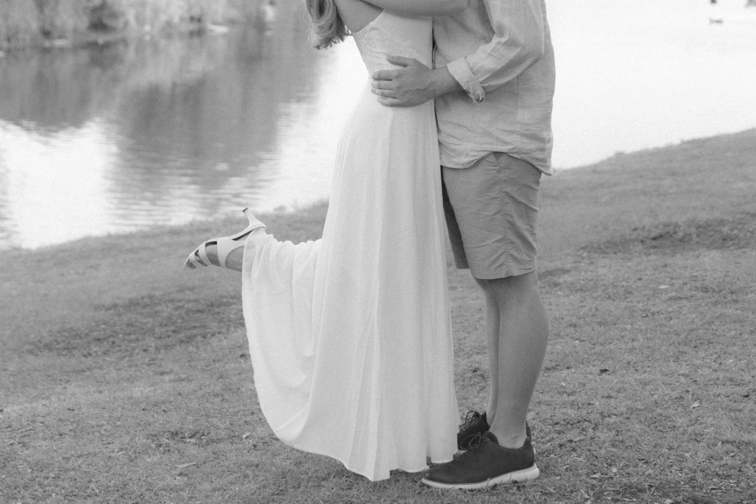 Black and white photo of a couple embracing near a lake, woman in flowing white dress, man in shorts and sneakers.
