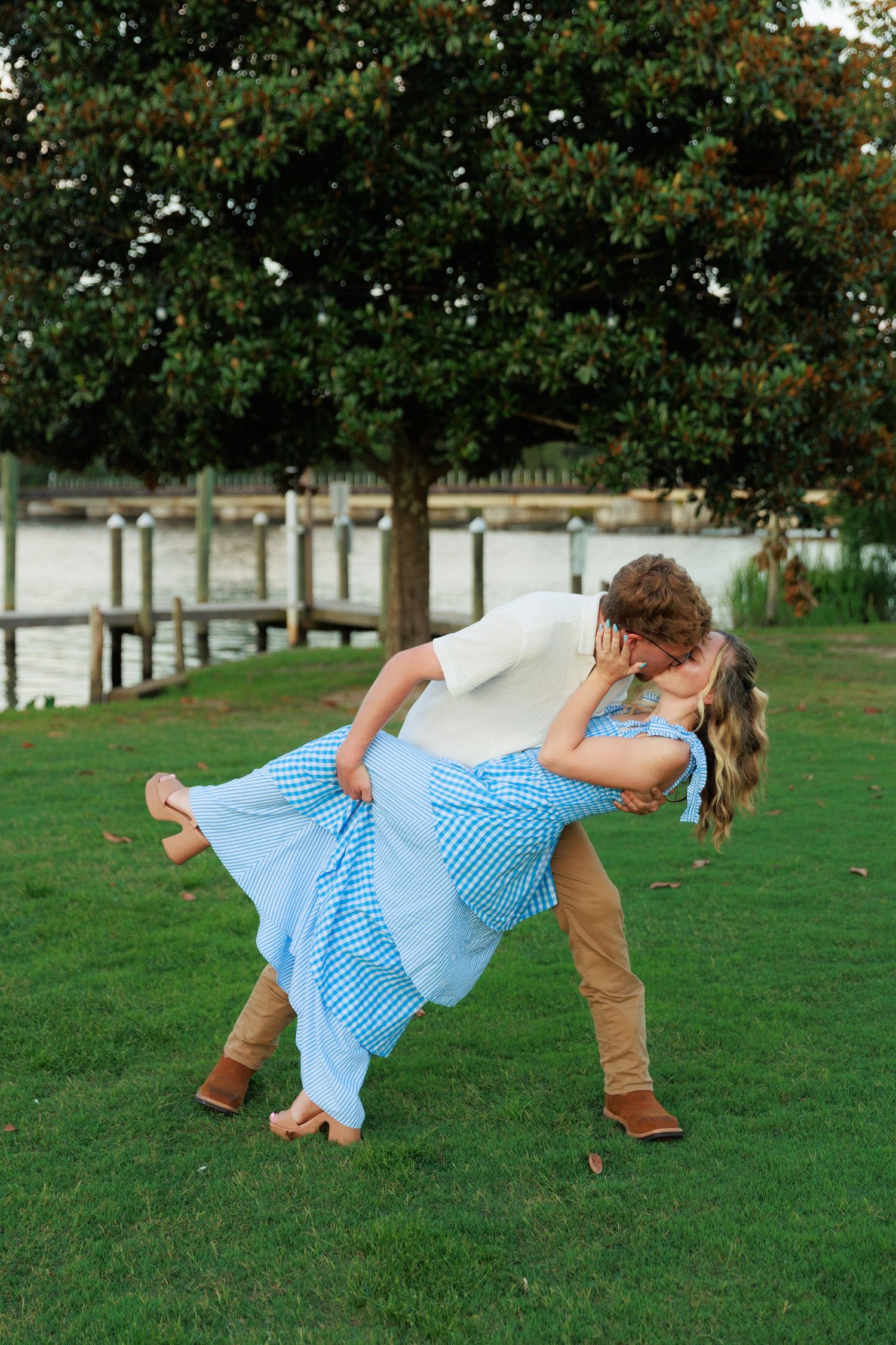 Couple sharing a romantic dip kiss on green grass near a waterfront dock, woman in blue gingham dress.