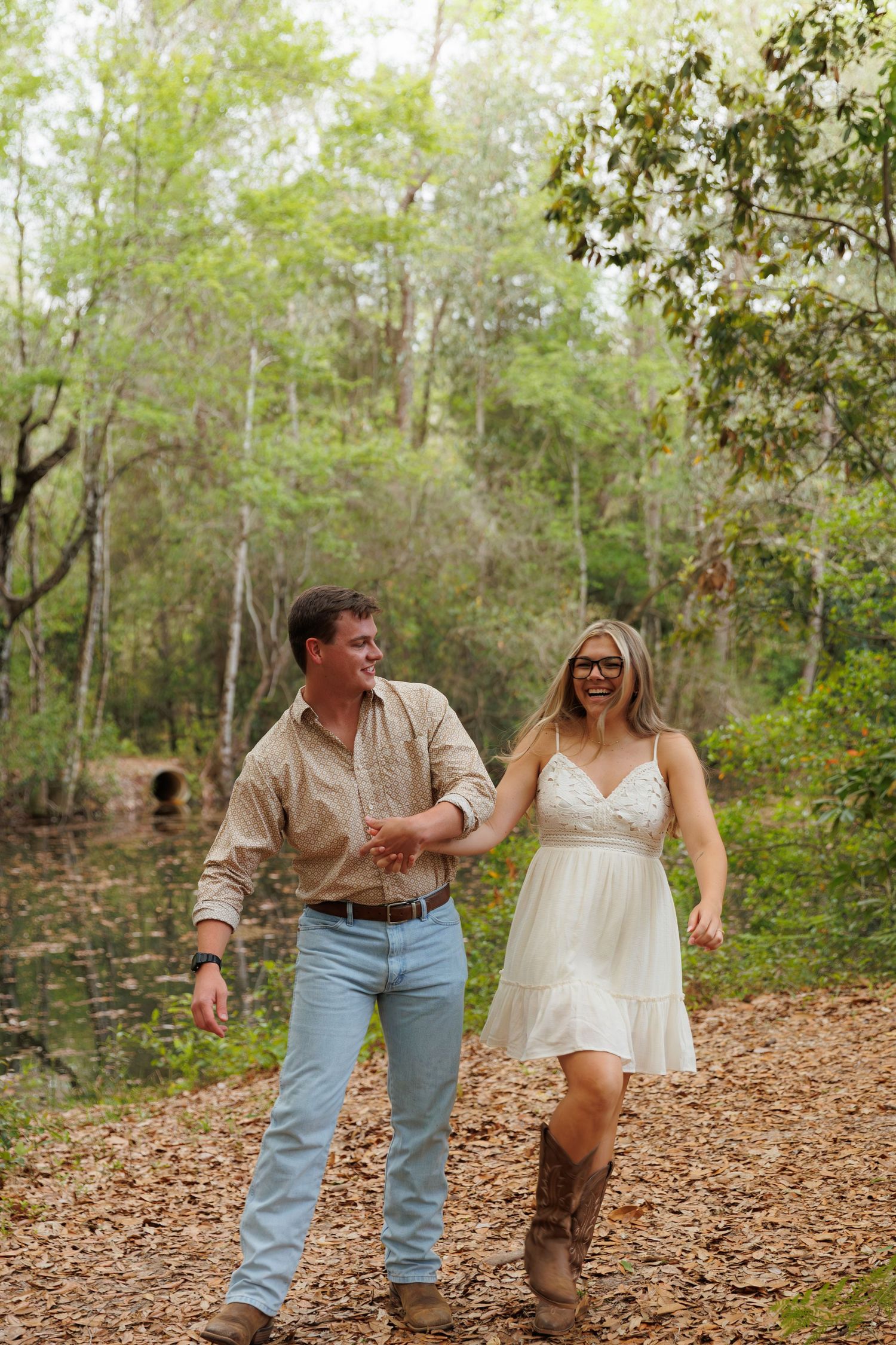 Happy couple holding hands and walking through a lush green forest path, woman in white dress and boots.
