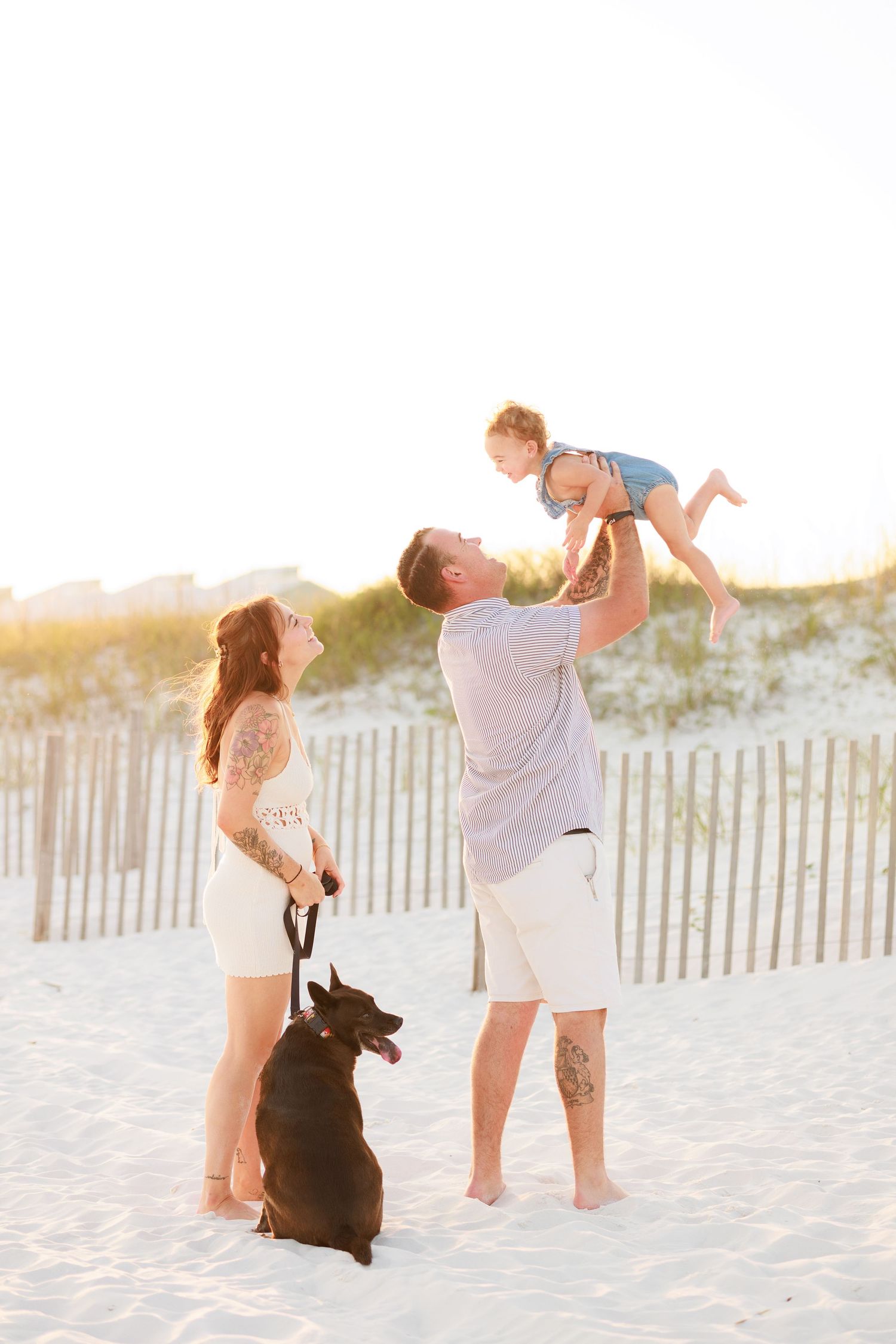 Family beach photo with parents lifting toddler in air while dark brown dog sits on white sand.