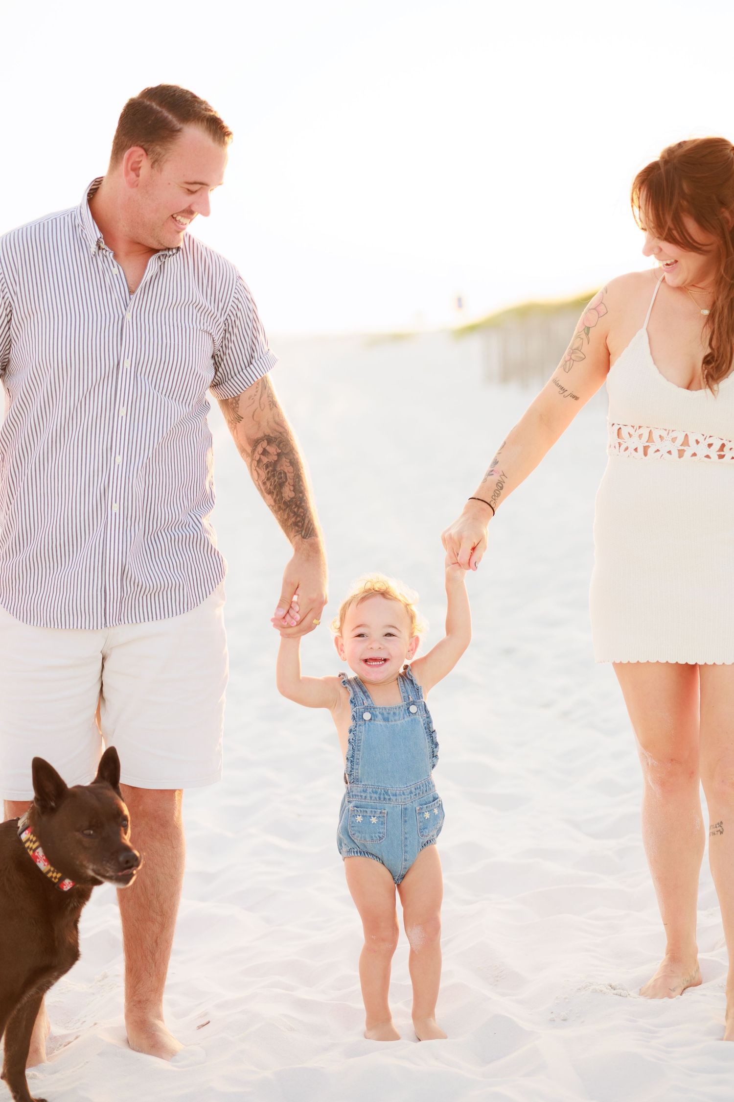 Smiling toddler in denim overalls holds parents hands on sunny beach with dog nearby.