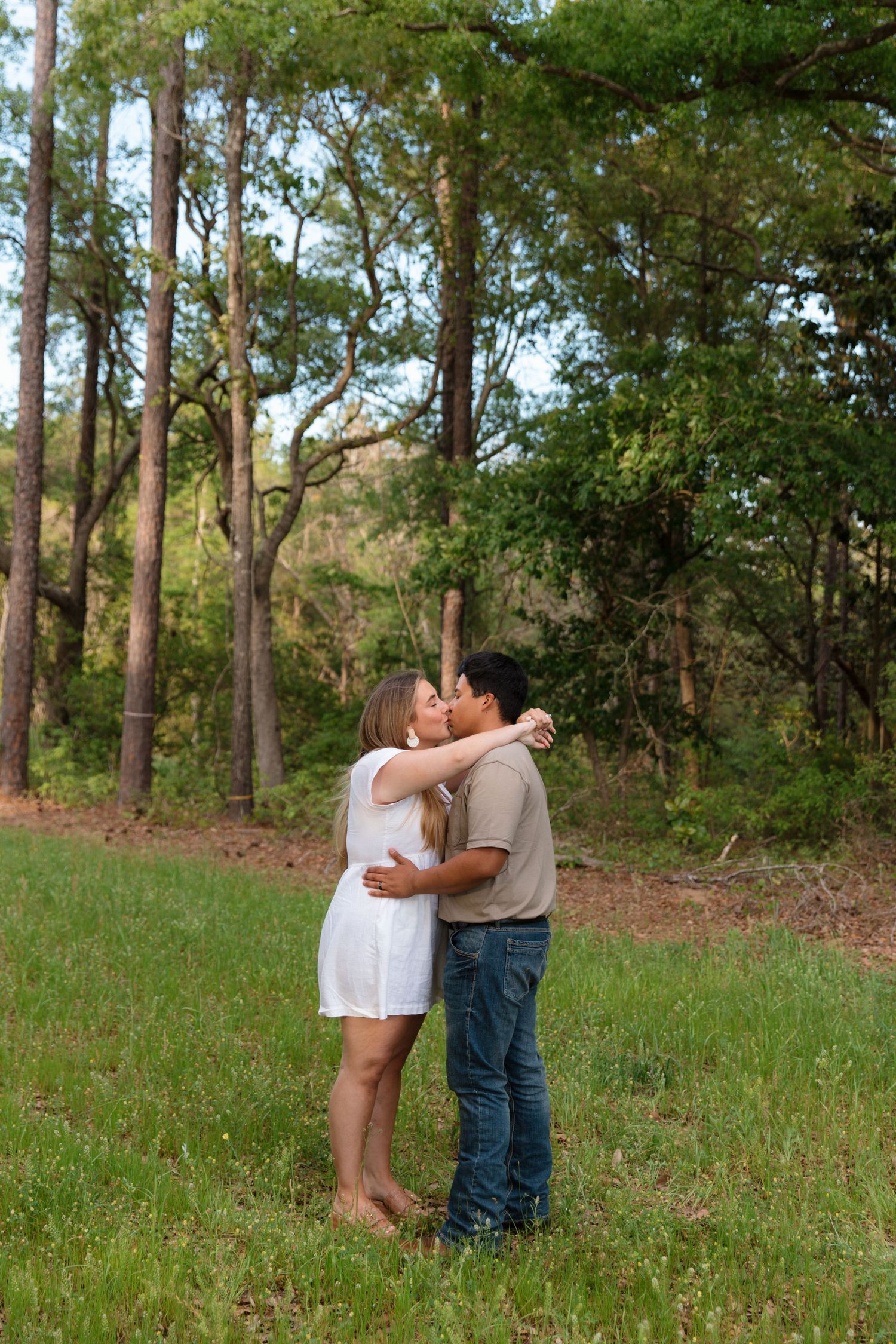 Couple sharing a romantic kiss in a lush green forest clearing, woman in white dress, man in jeans and tan shirt.