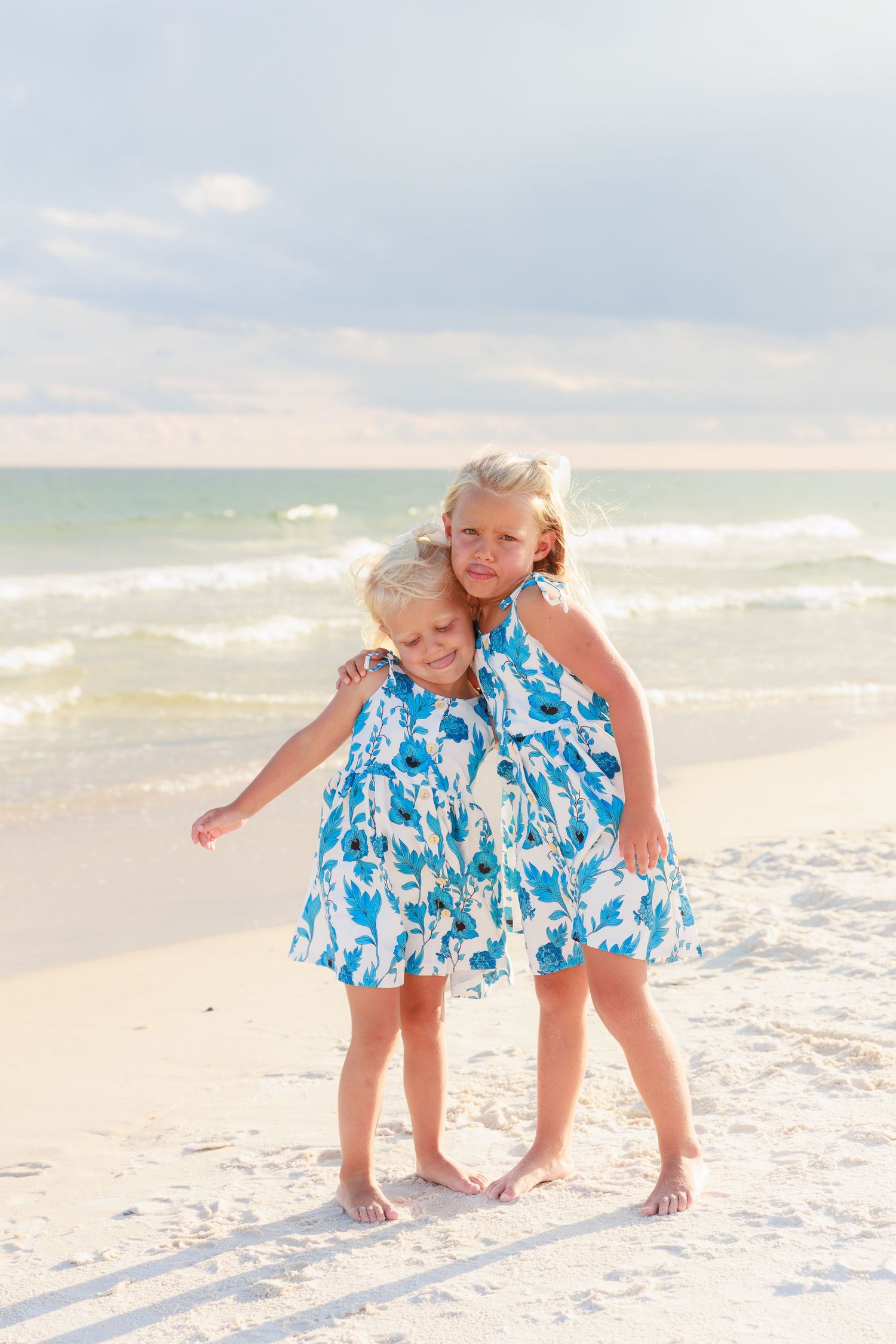 Two young blonde sisters in matching blue floral dresses hugging on a sandy beach with ocean waves behind them.
