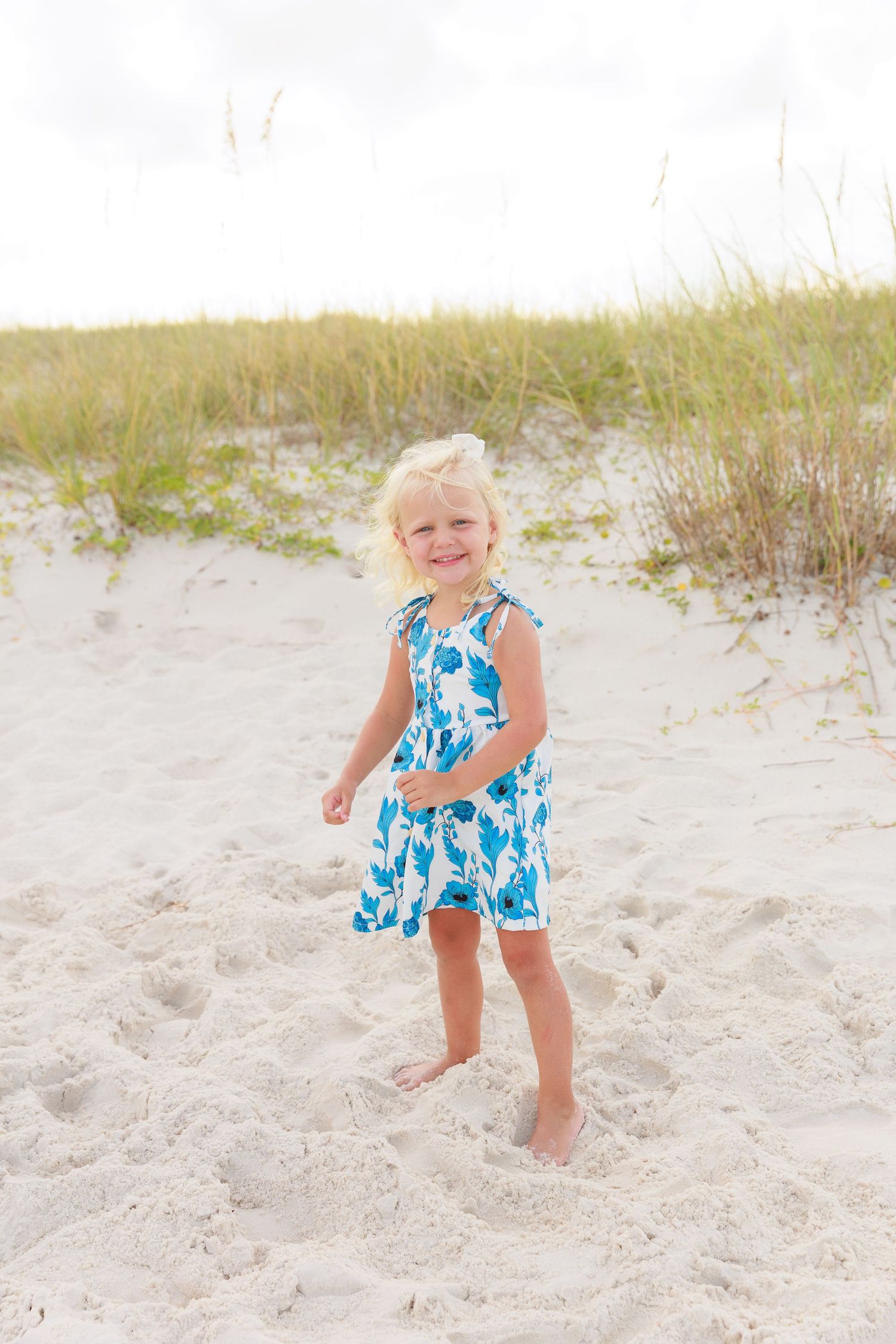 A happy blonde girl in a blue floral sundress stands barefoot on white sand at the beach.