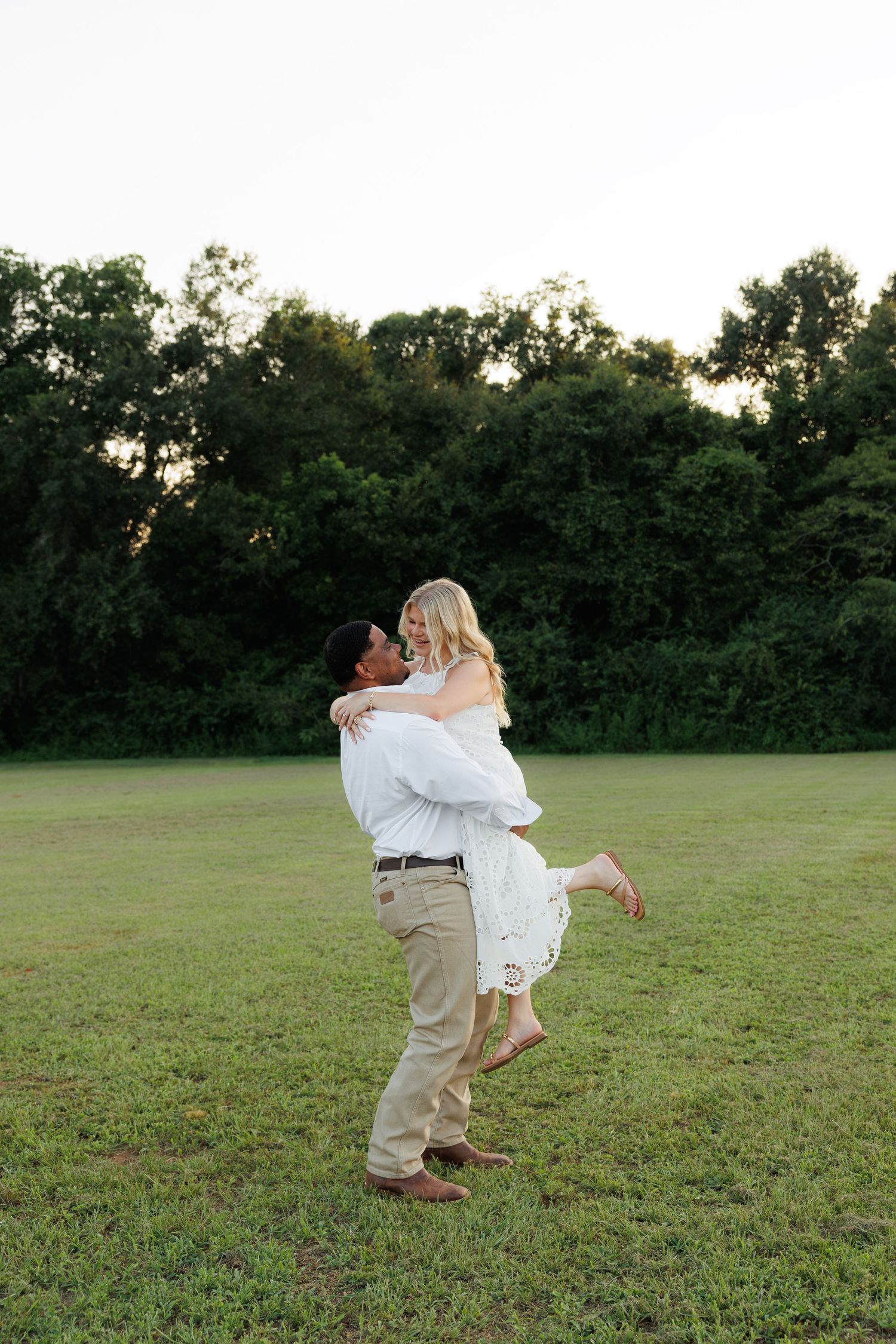 A man lifts a woman in a white dress in a green field at sunset, sharing a romantic moment outdoors.