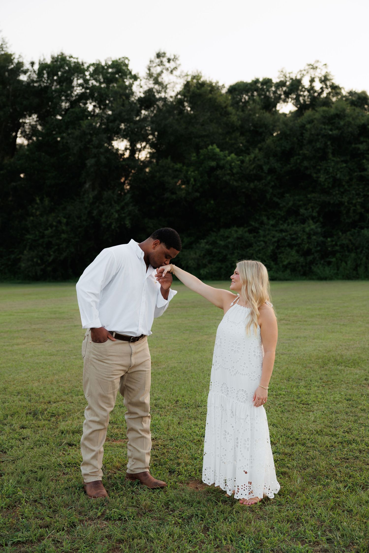 Man in white shirt kisses womans hand in green field at dusk, woman wearing elegant white lace maxi dress.