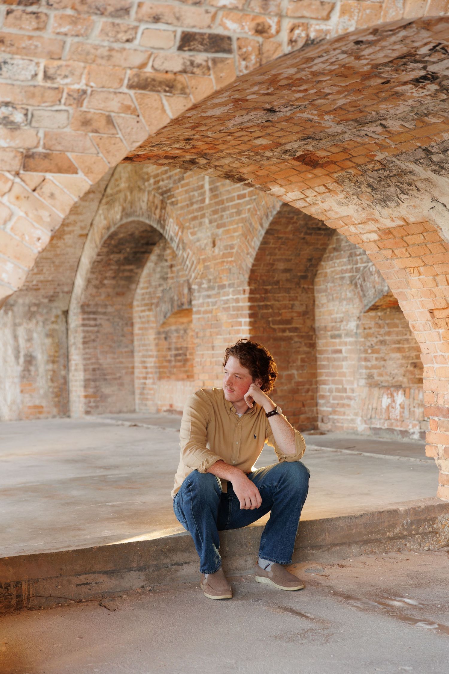 Young man in tan shirt sitting under historic brick arched corridor in contemplative pose.