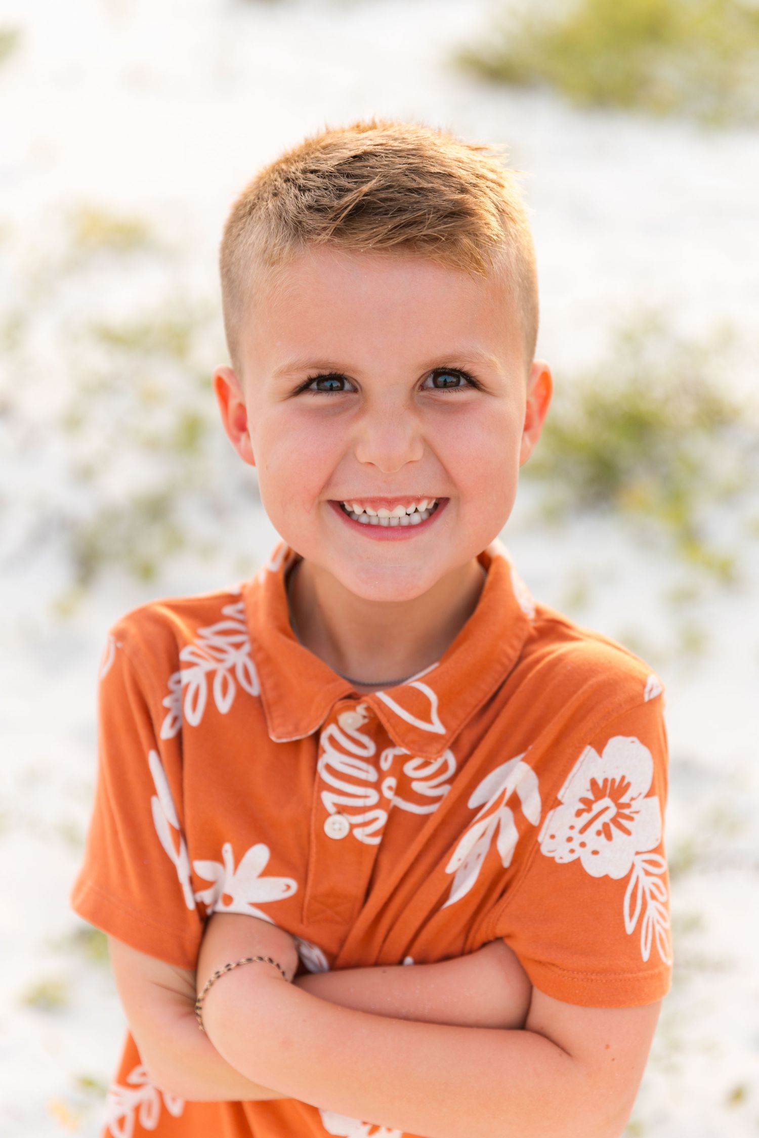 Young child wearing an orange floral shirt poses on the beach with a bright smile.