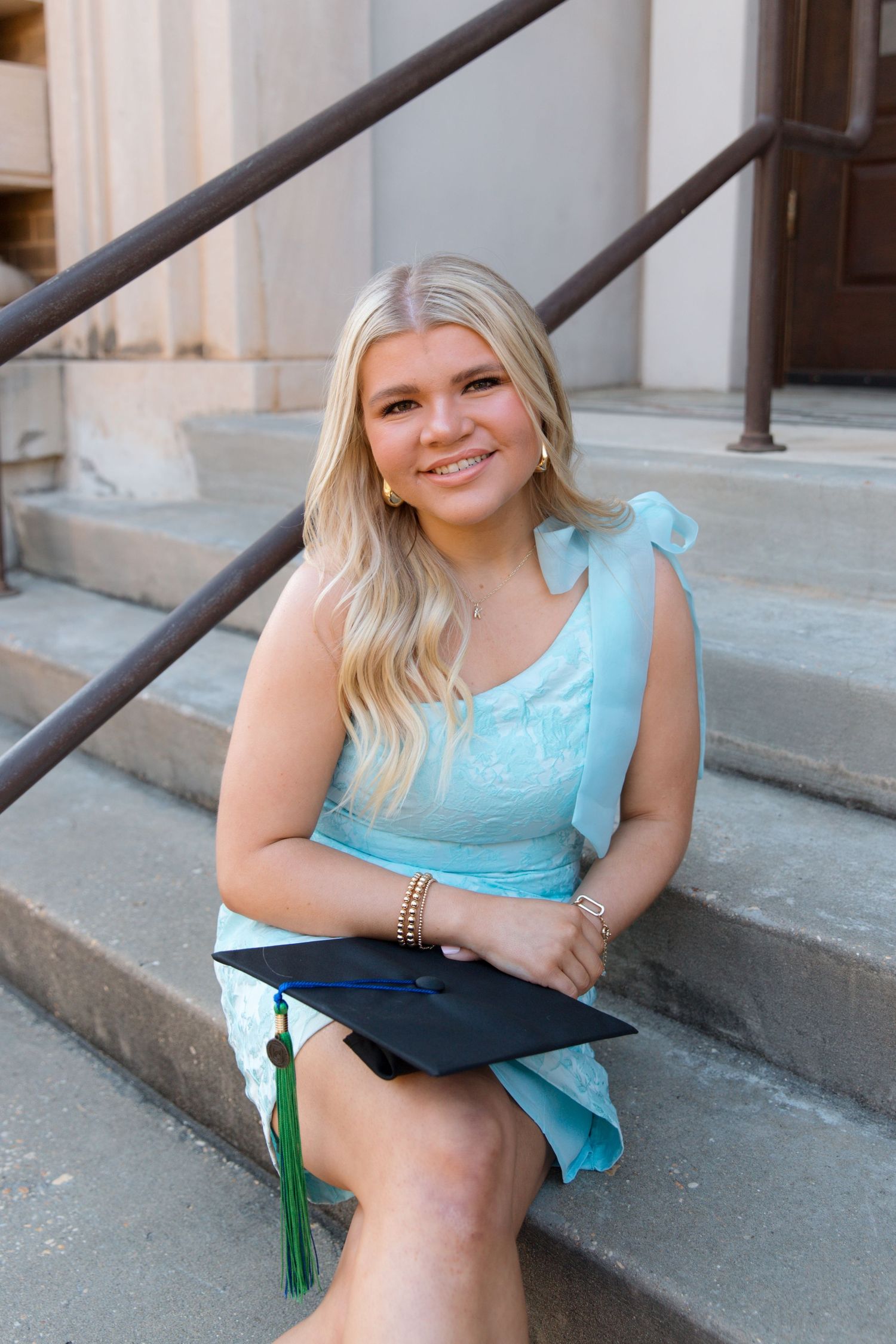 Graduate in a turquoise dress poses on stone steps with a graduation cap and green tassel.