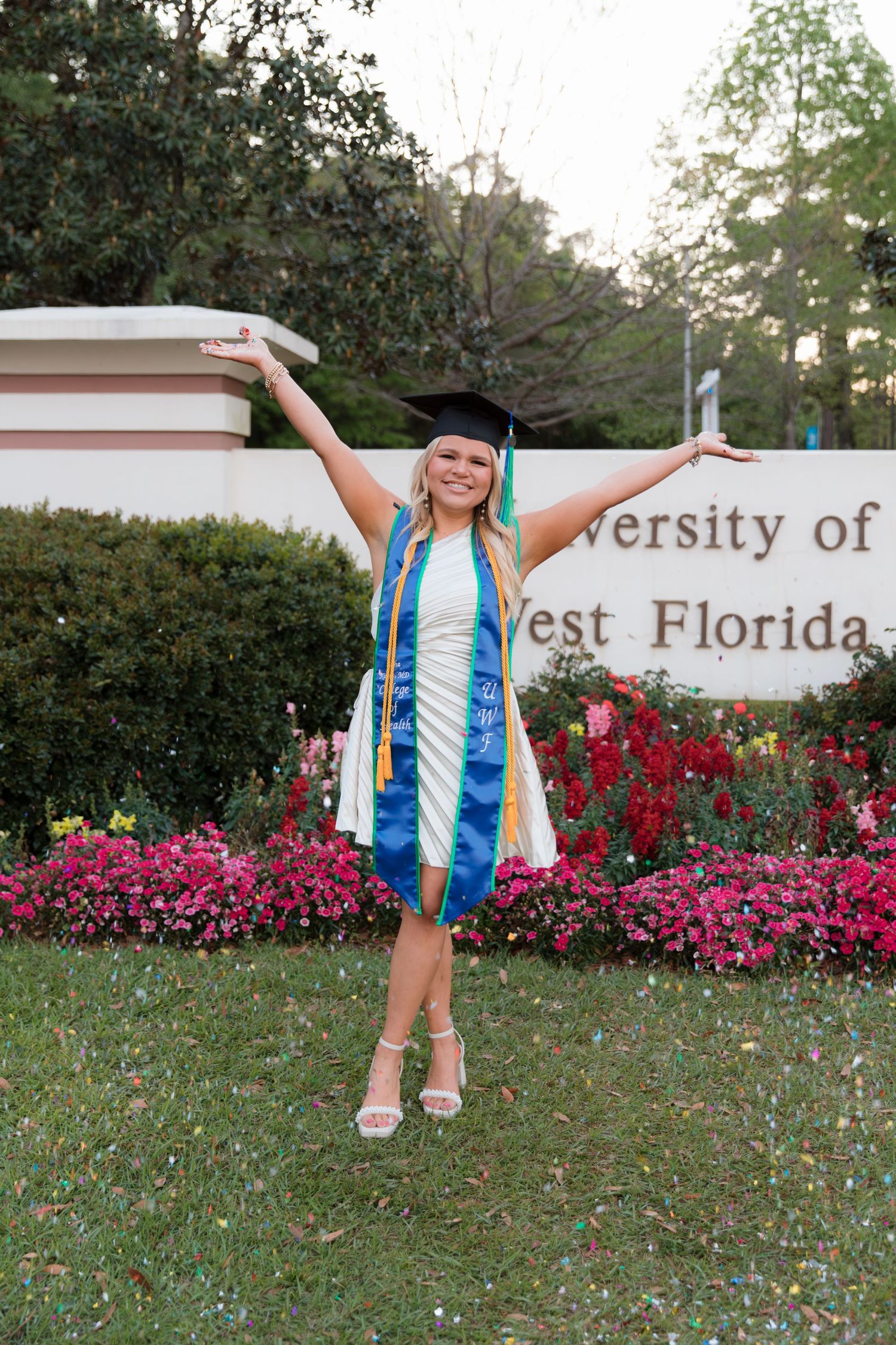 A graduate poses with arms raised in celebration near a university sign surrounded by vibrant pink flowers.