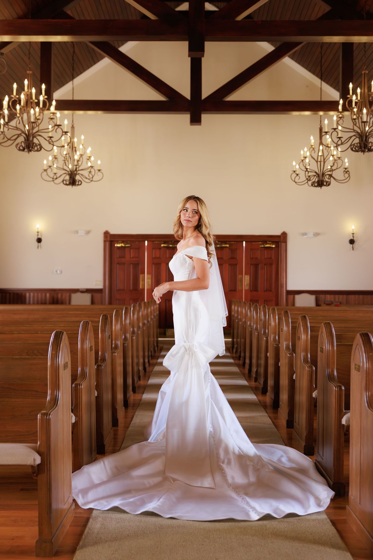 A white wedding dress flows elegantly across wooden church pews in a chapel with exposed beam ceilings and chandeliers.