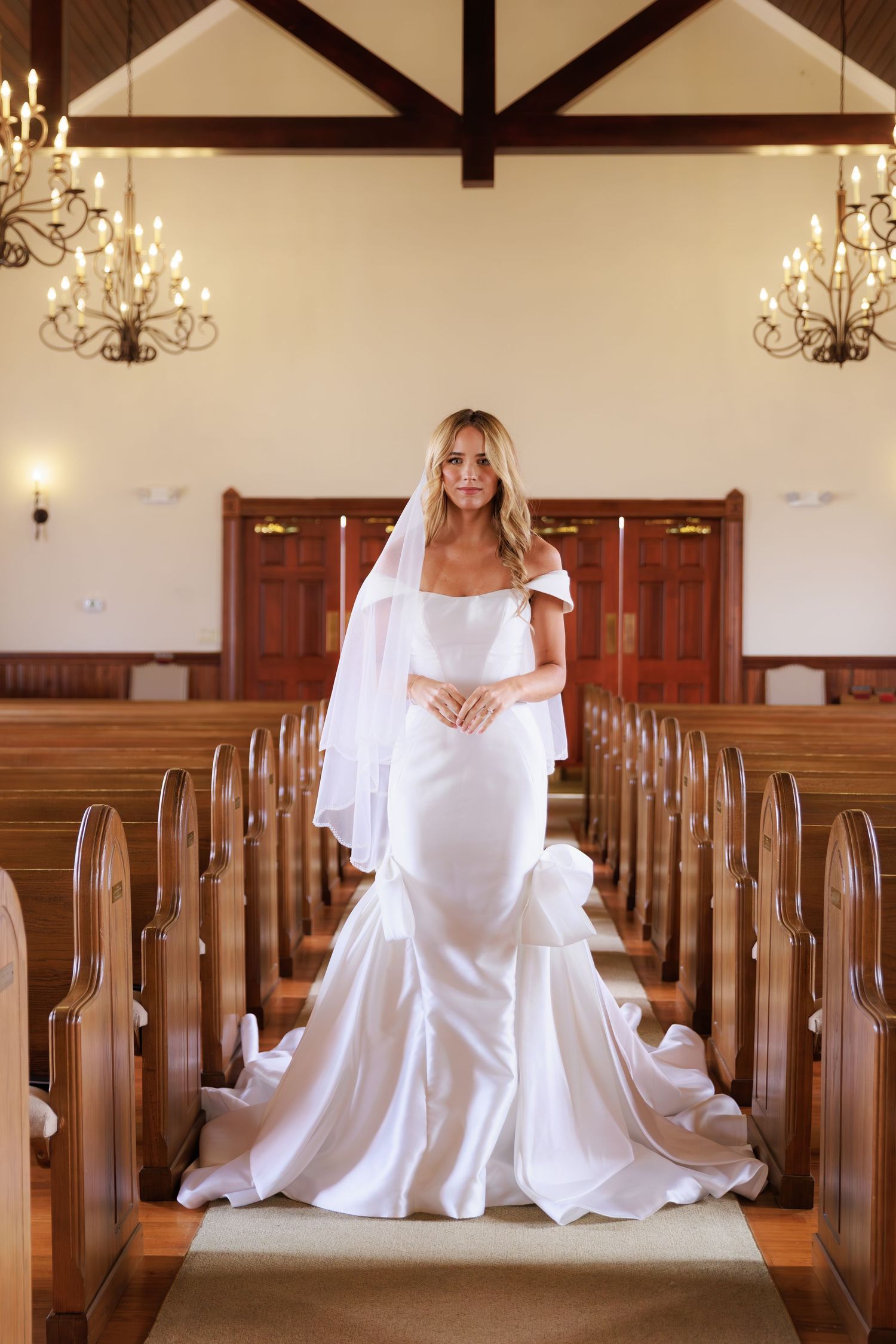 A person in a flowing white dress sits gracefully in a wooden church pew with chandeliers overhead in a chapel setting.