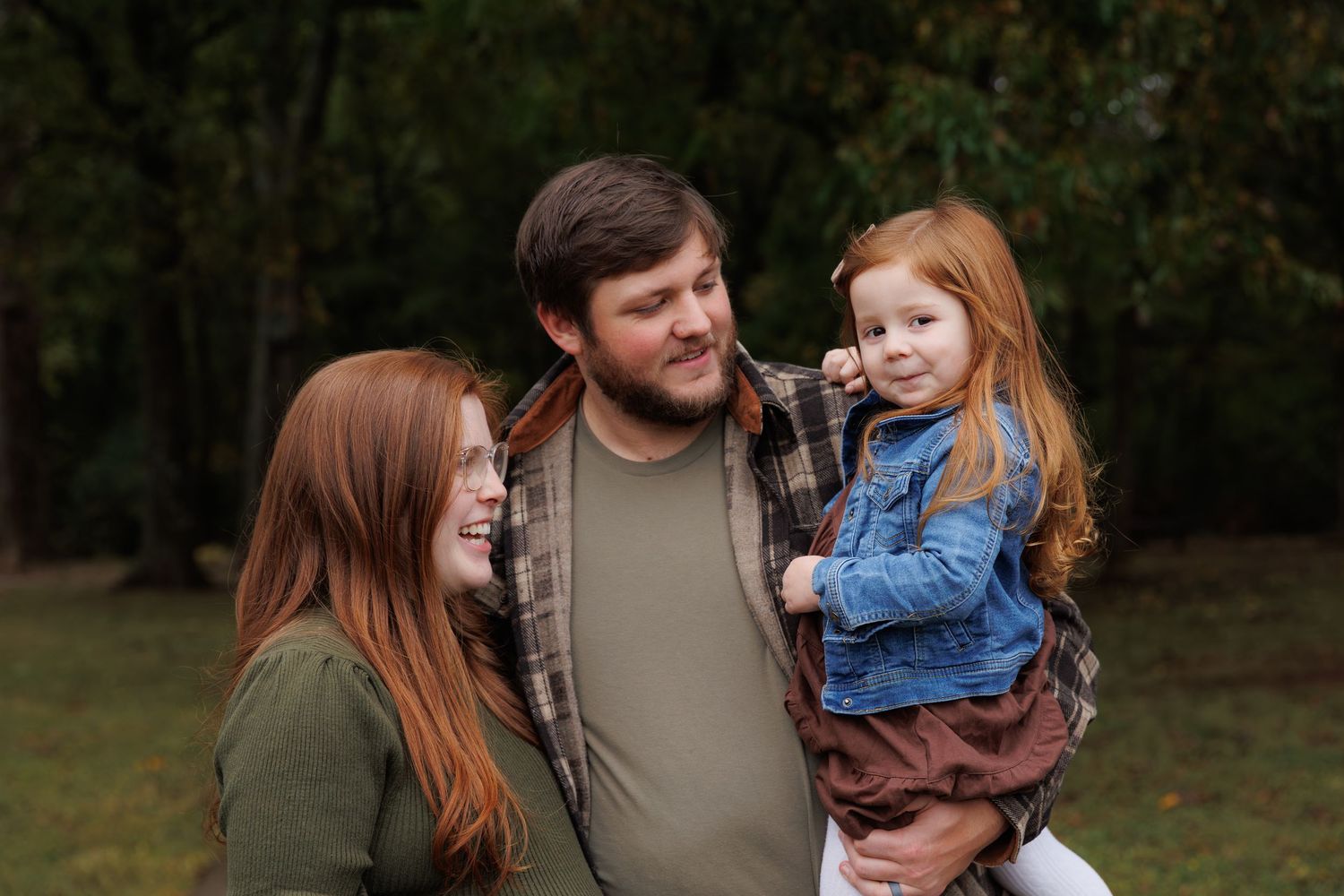 A family in casual fall attire stands together outdoors against a wooded backdrop during an autumn photo session.