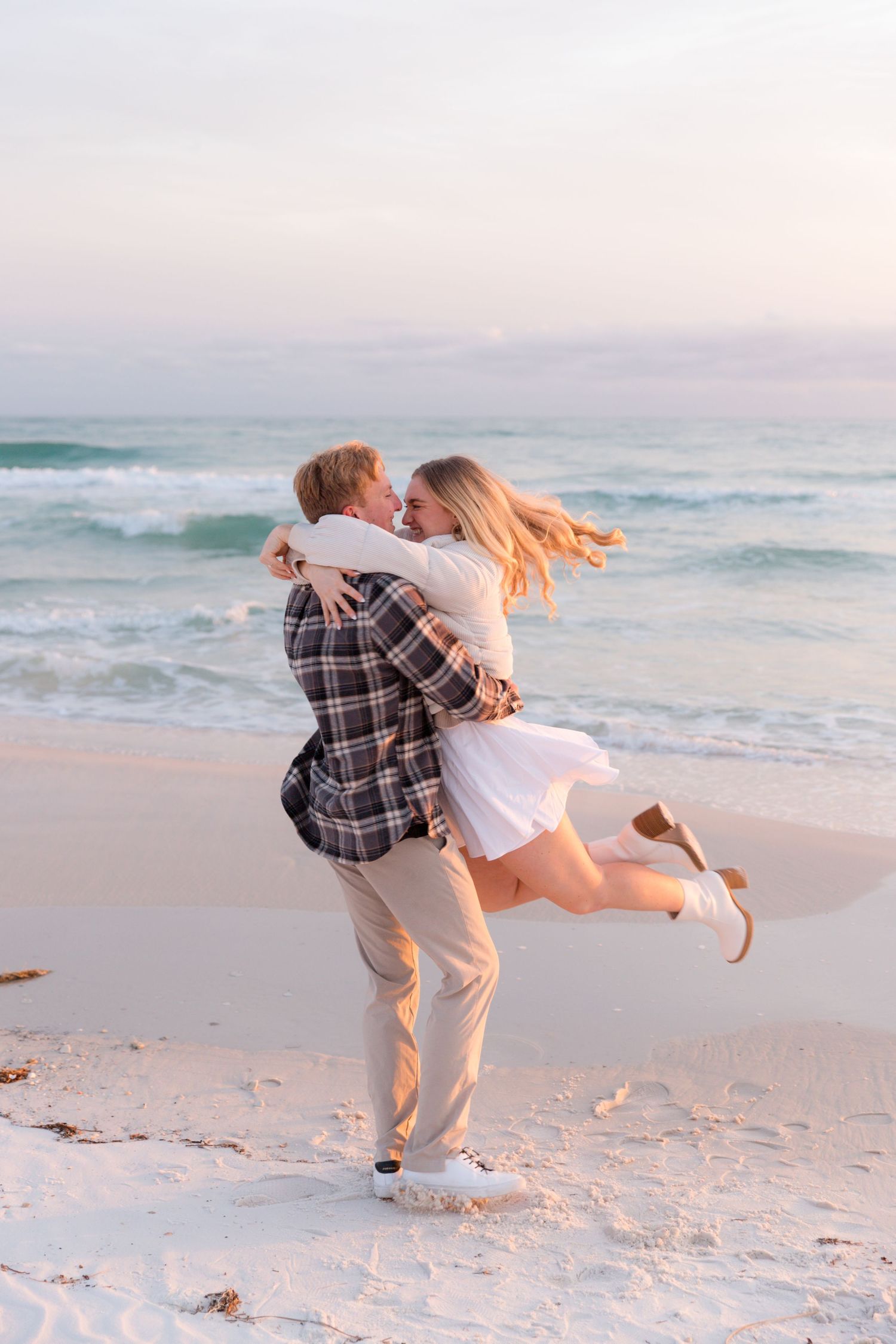 A couple embraces and spins playfully on a sandy beach at sunset with ocean waves in the background.