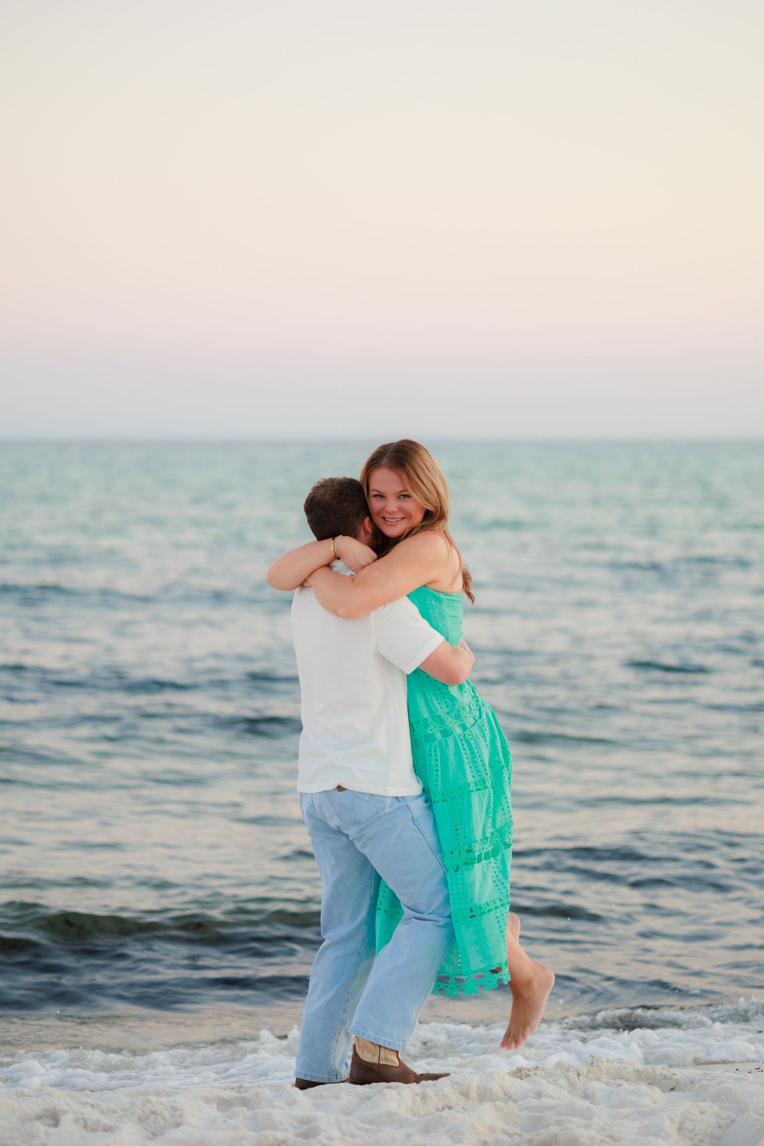 A couple embraces on the beach at sunset with turquoise water and soft pink sky in the background.
