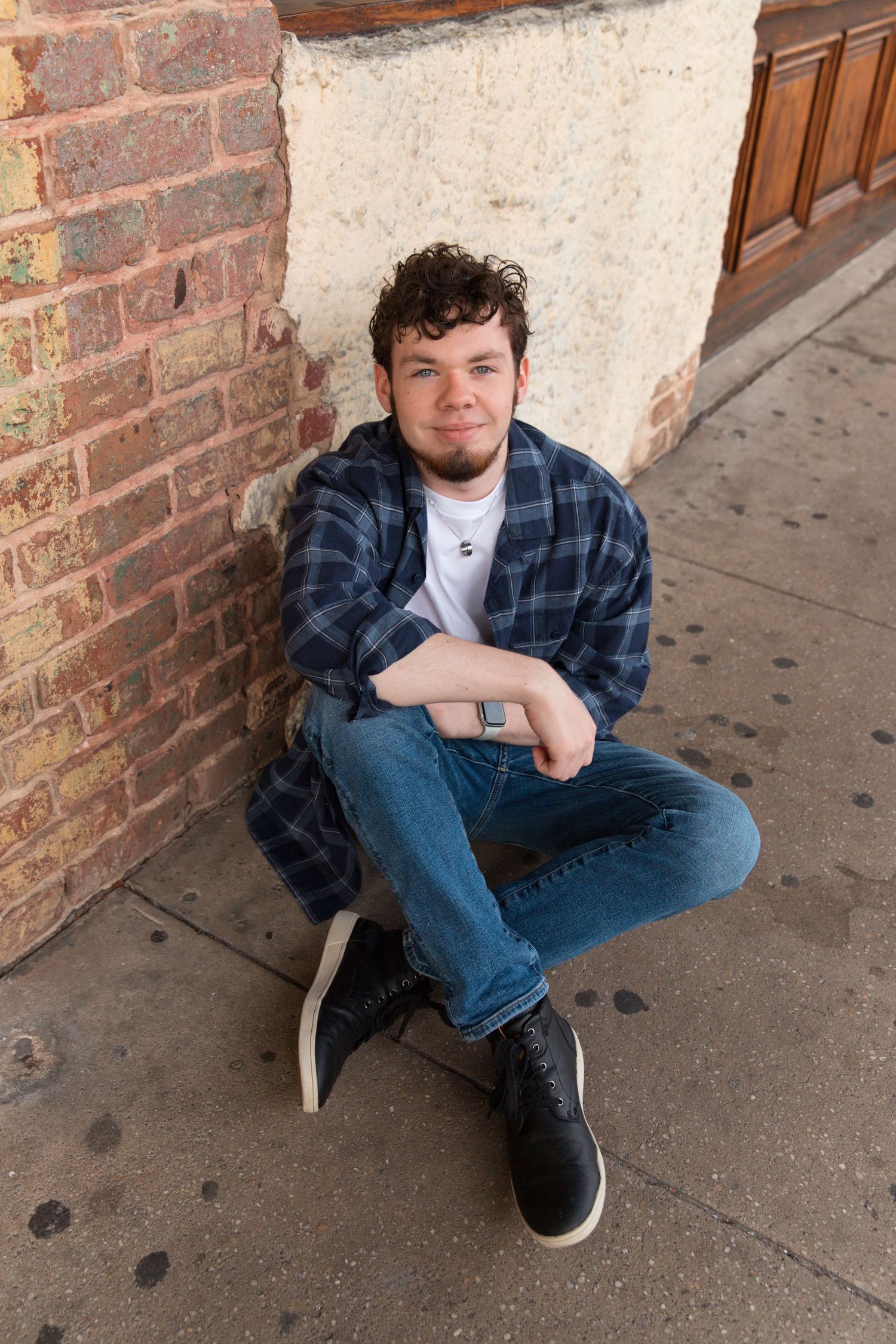 Relaxed casual pose sitting on sidewalk against brick wall in plaid shirt and jeans.