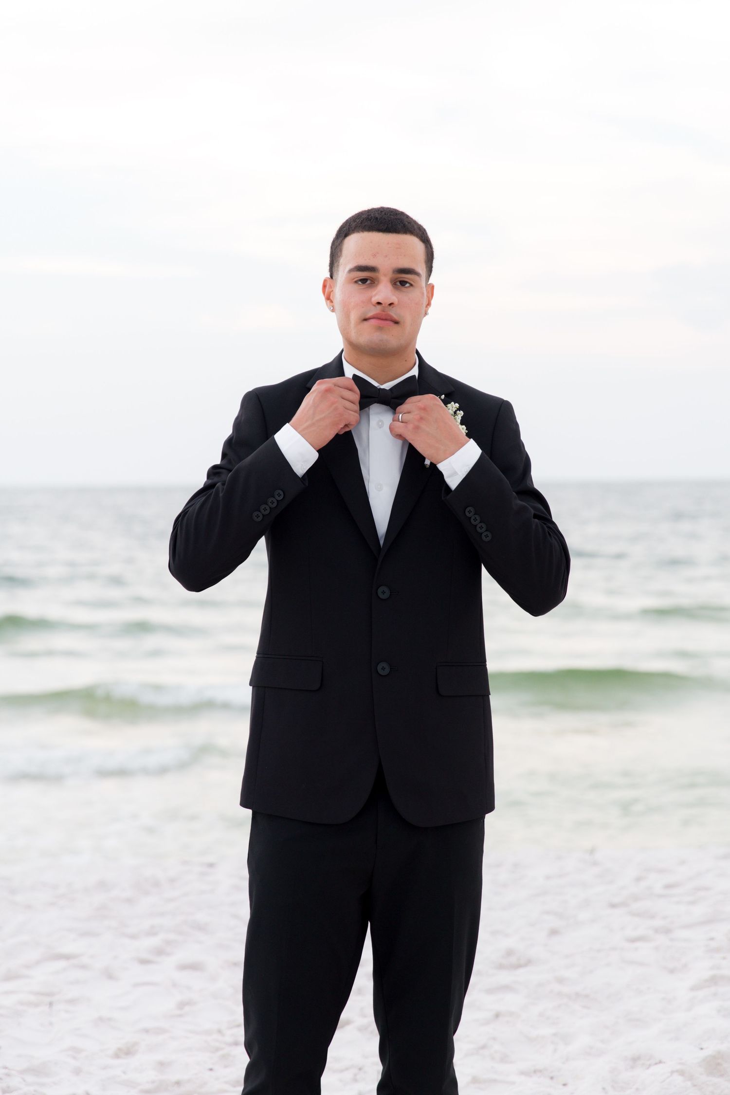 Groom adjusting bowtie in classic black tuxedo on beach with ocean waves in background.
