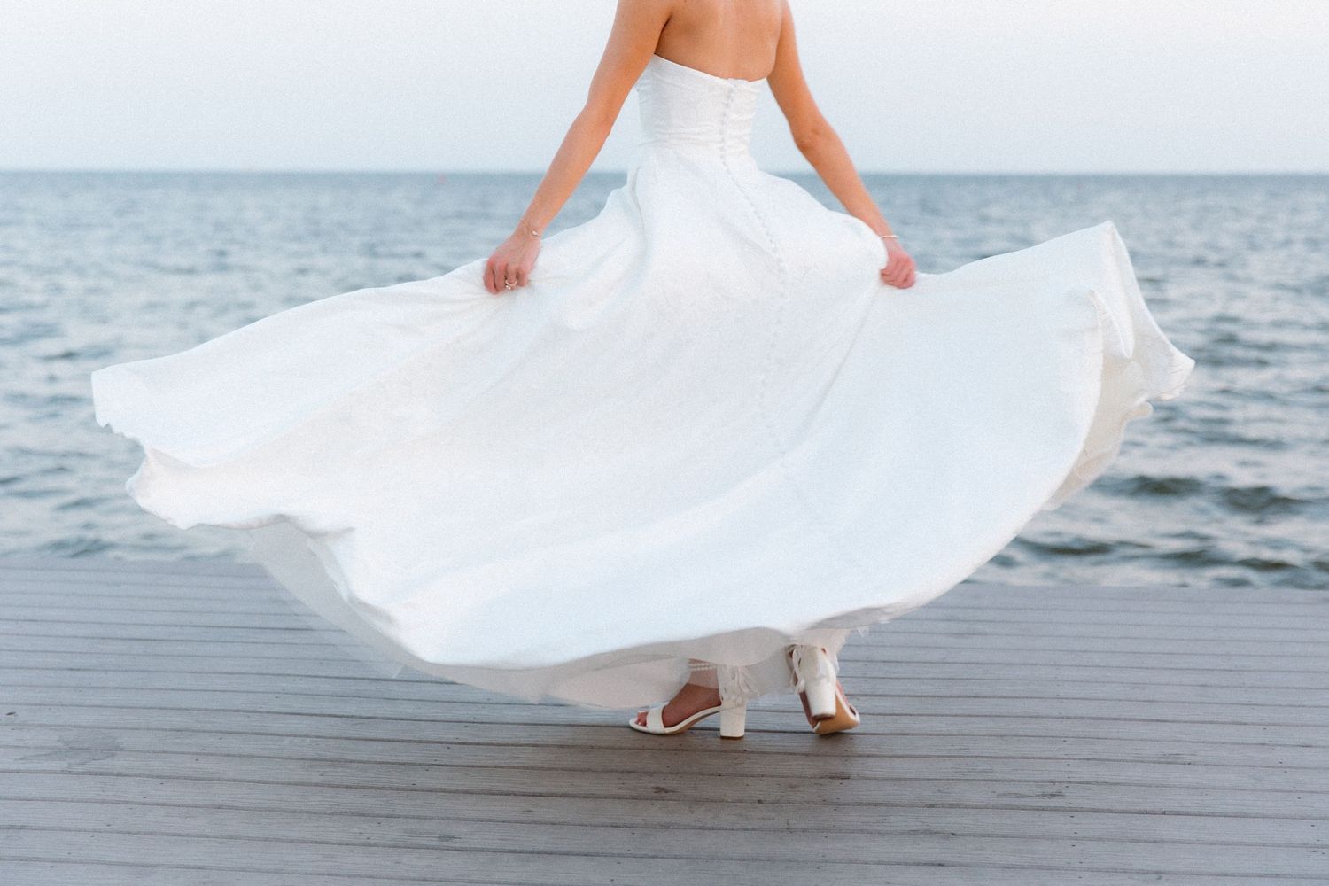 Elegant bride spins in white dress on wooden dock with ocean view.