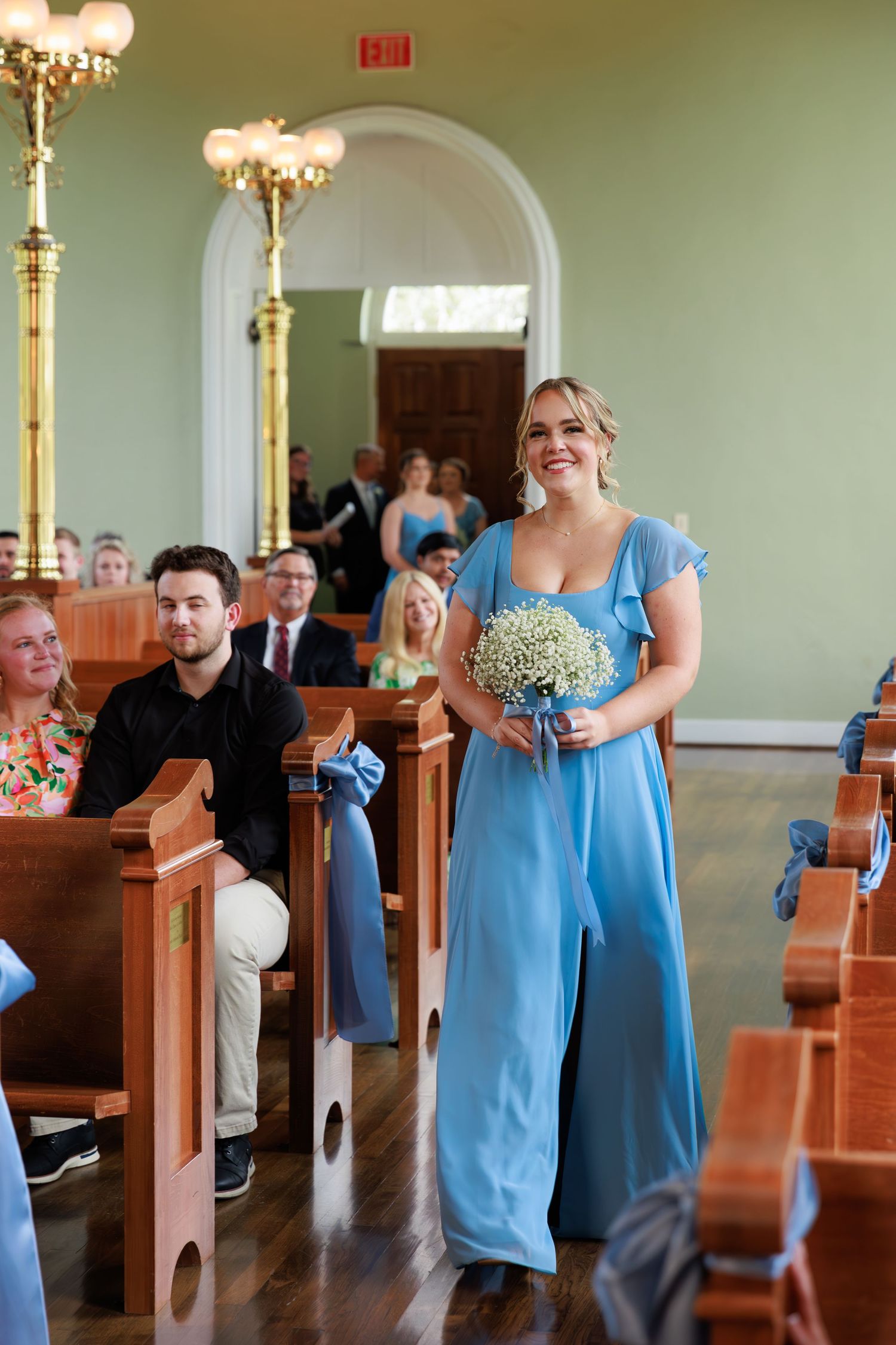 Bridesmaids in light blue dresses walk down church aisle carrying bouquets.
