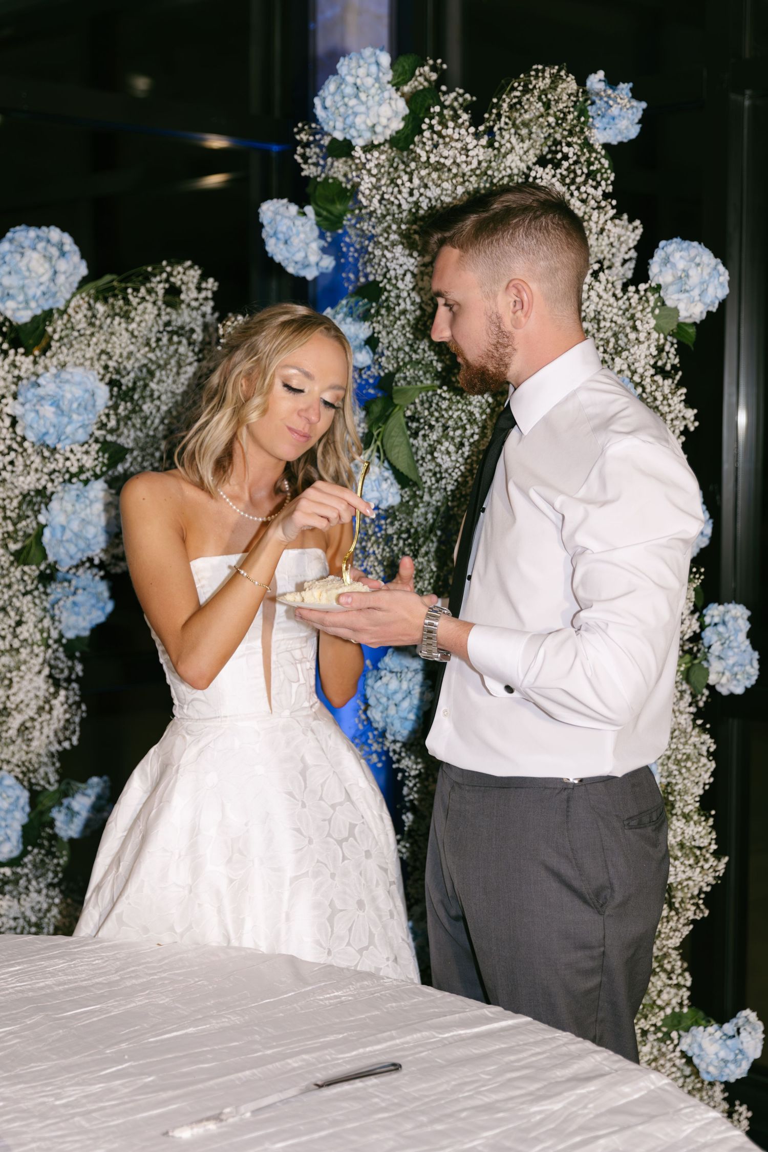 A couple shares wedding cake beneath an arch decorated with blue hydrangeas and baby's breath flowers.