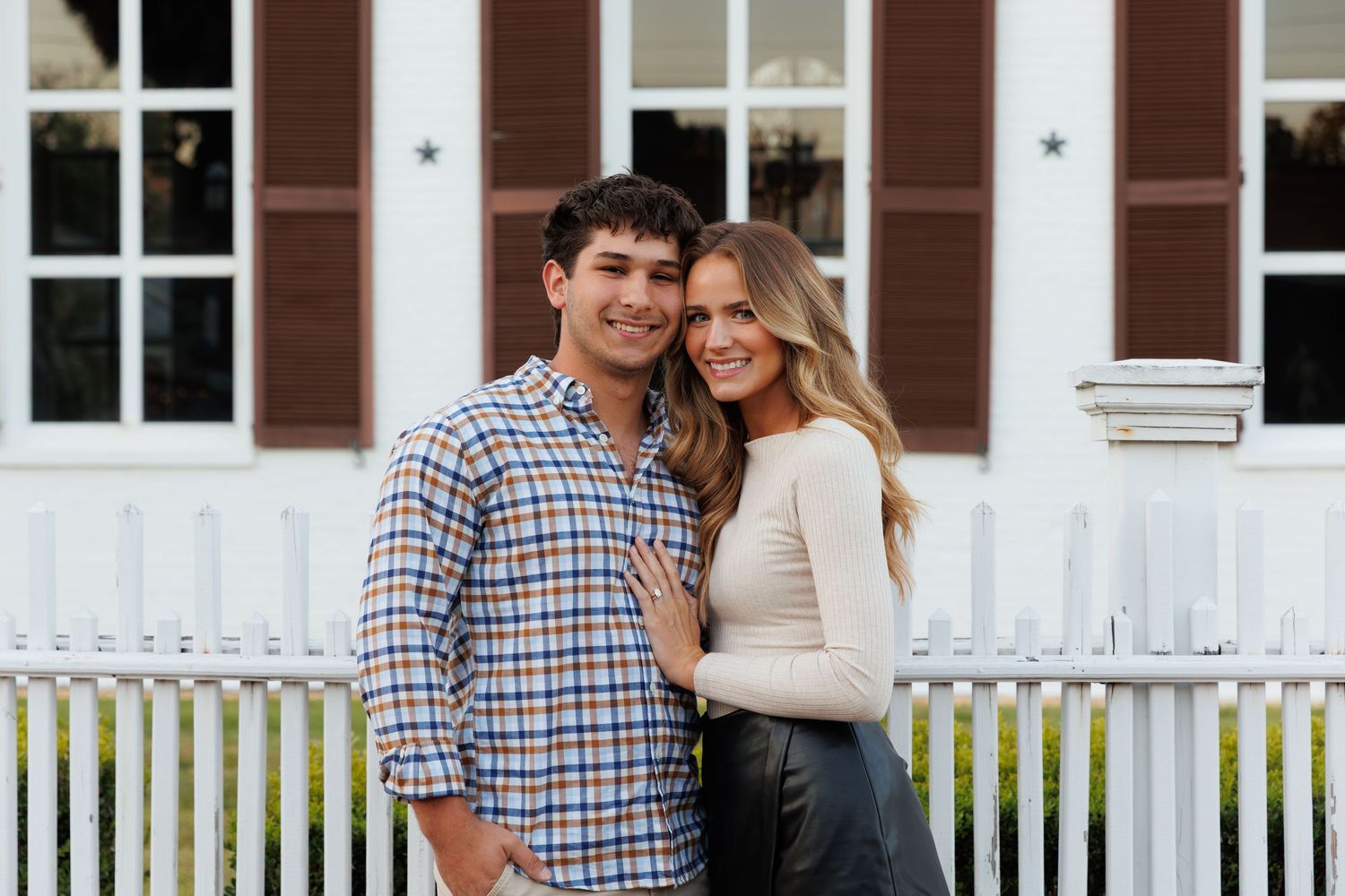 Young couple smiling together in front of white picket fence and historic building with brown shutters.