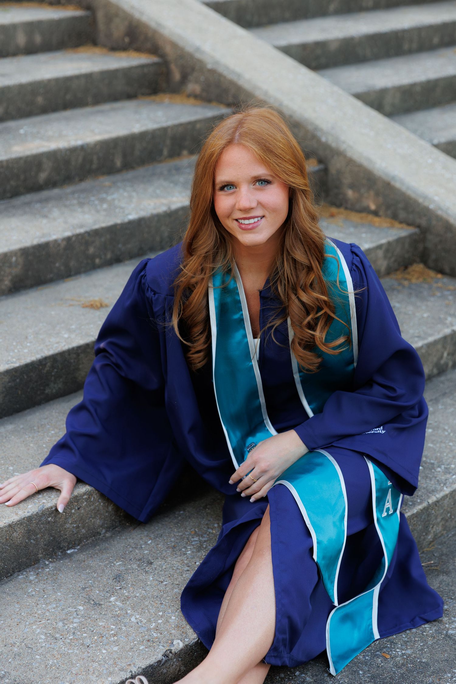 Smiling redhead woman in blue graduation gown with teal sash sitting on stone steps outdoors.