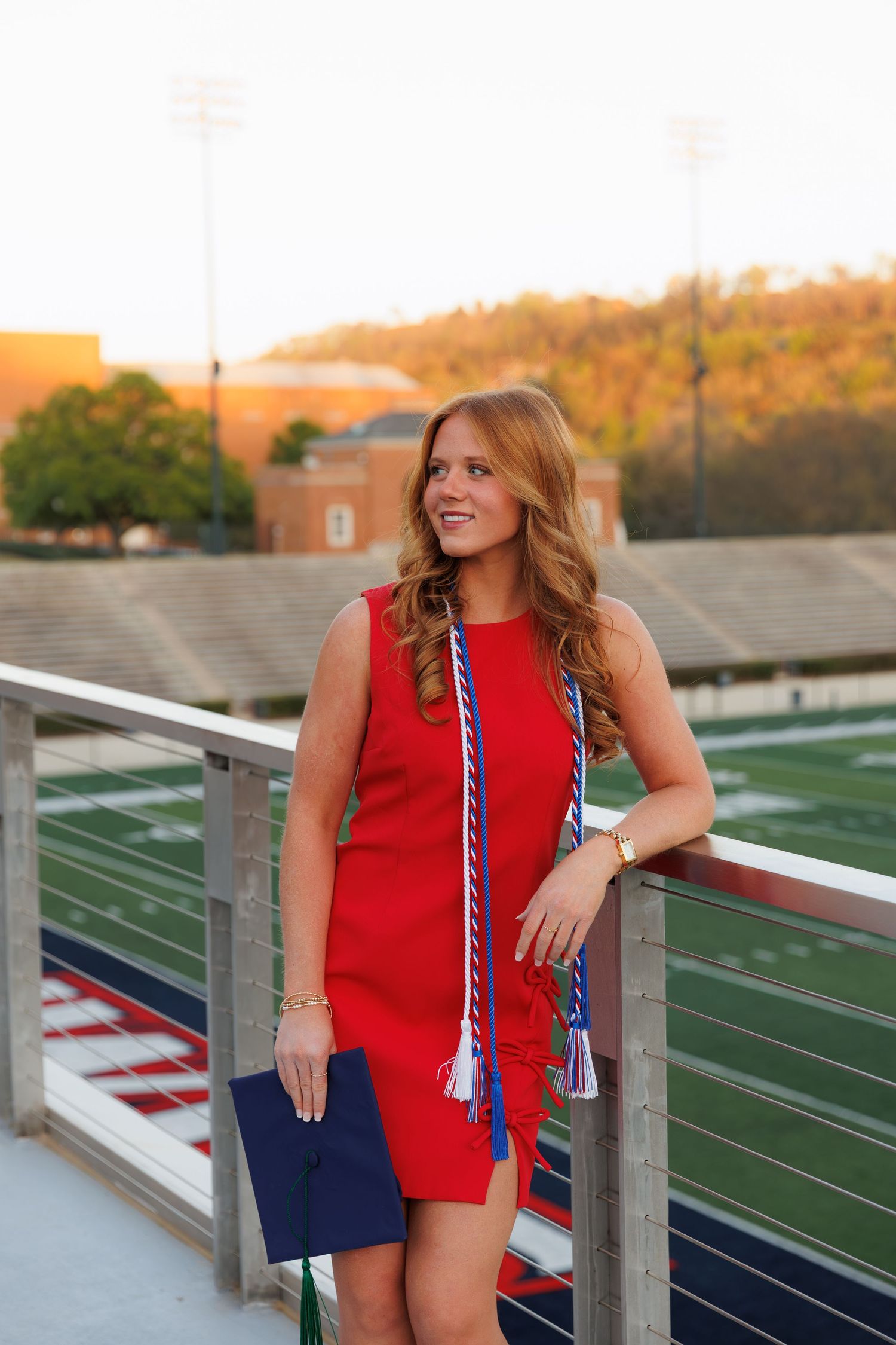 College graduate in red dress with honor cords gazes away on sunny stadium balcony.