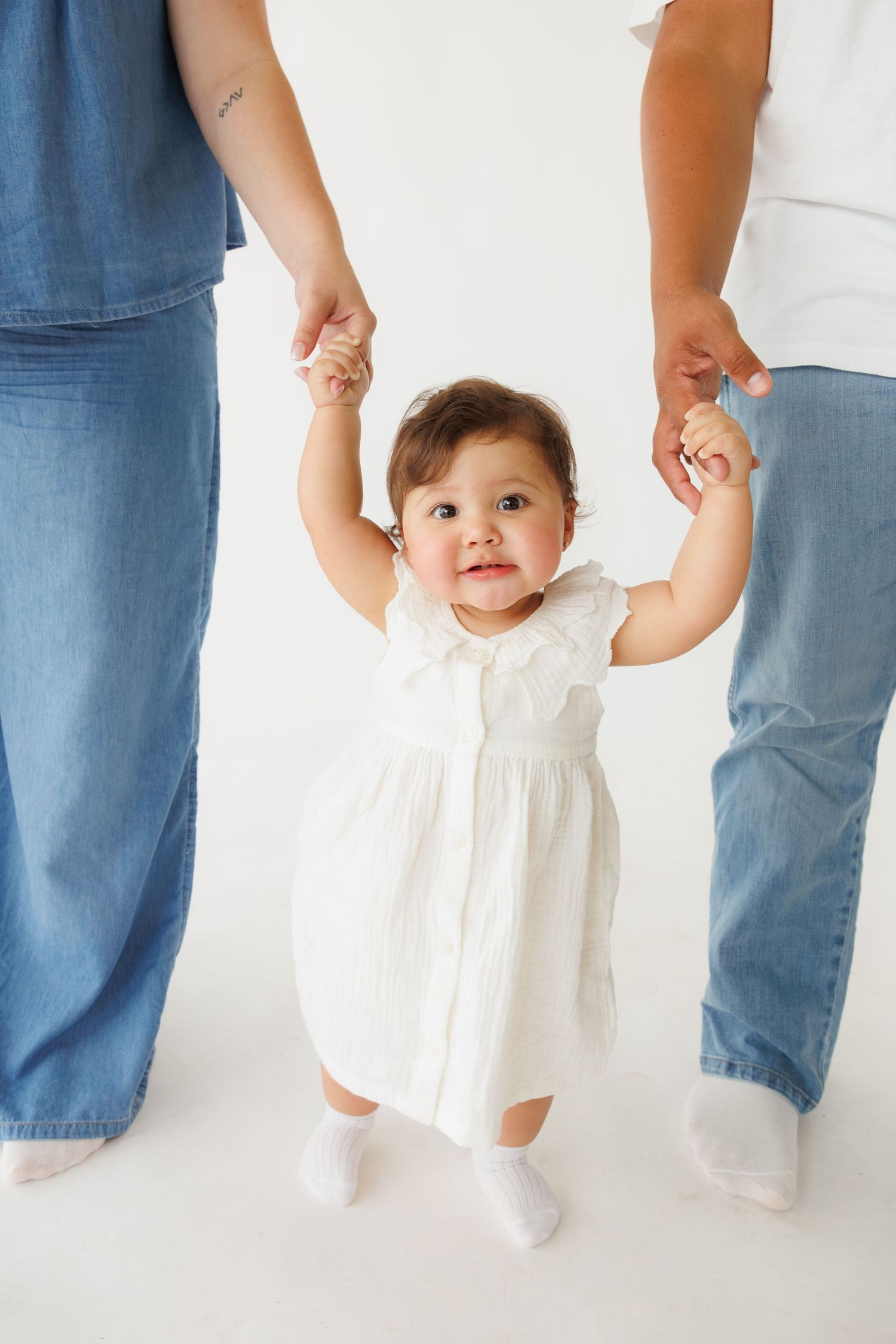 Smiling baby girl in white dress taking first steps while holding hands with two parents in jeans on white background.