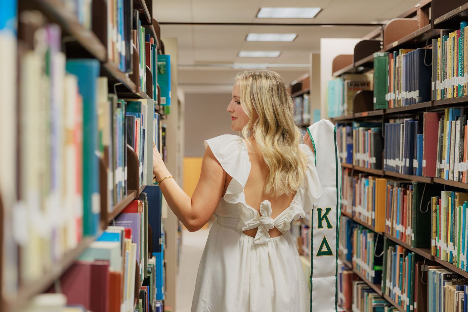 Blonde woman in white Kappa Delta dress browsing books in a college library aisle.