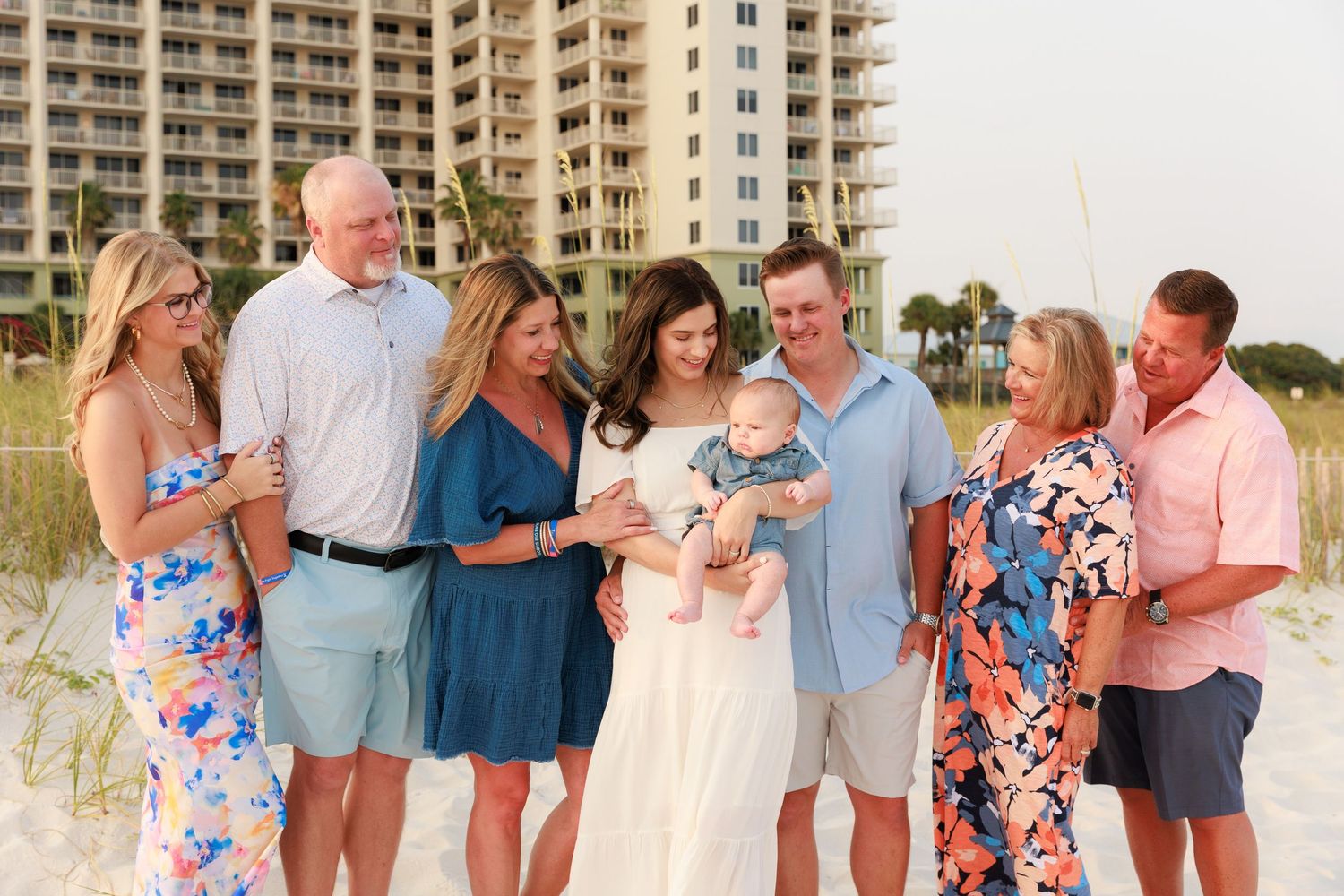 Large family group photo on sandy beach with high-rise condos in background, celebrating new baby.