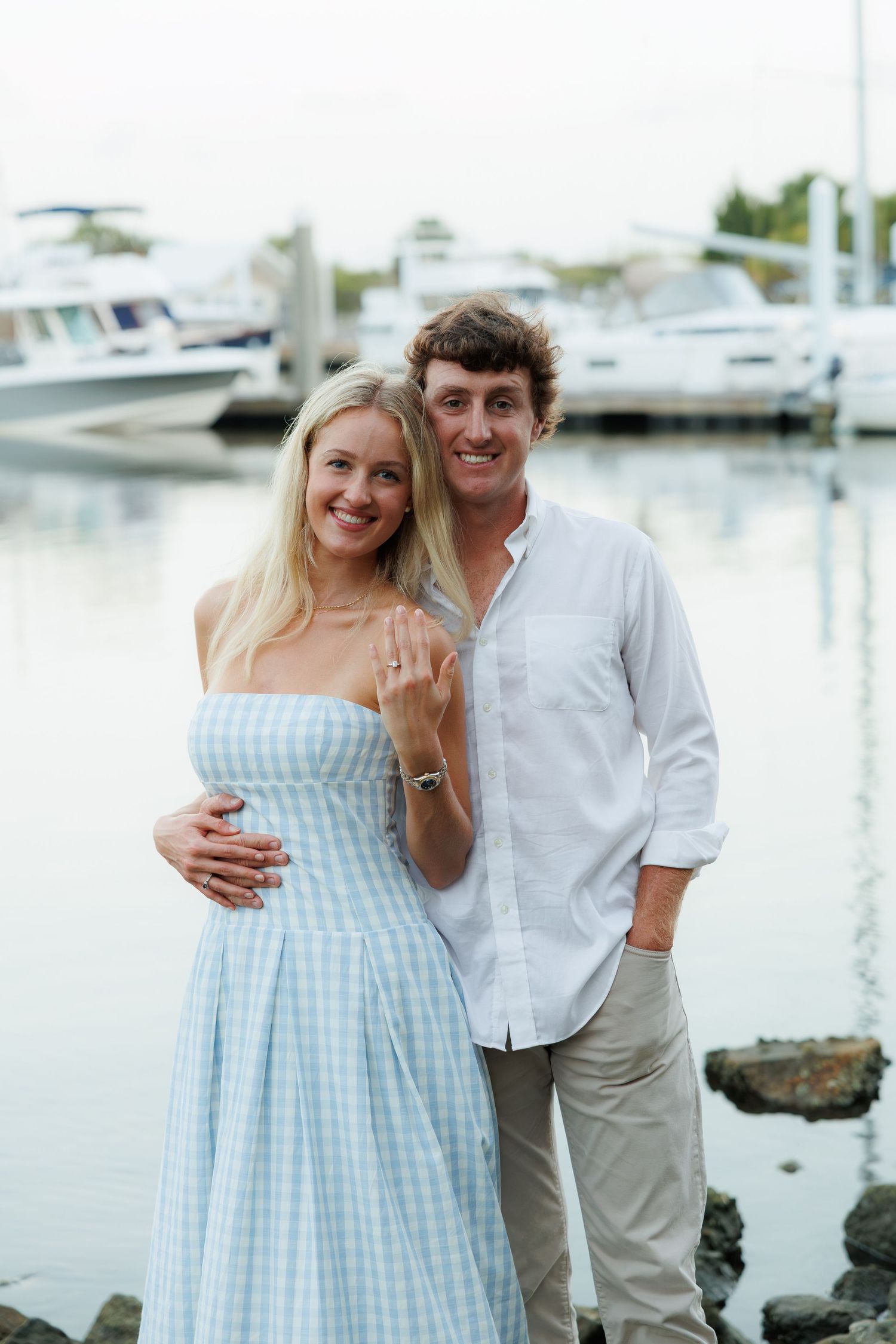 Engaged couple showing off ring near marina, woman in blue gingham dress, man in white shirt, boats in background.