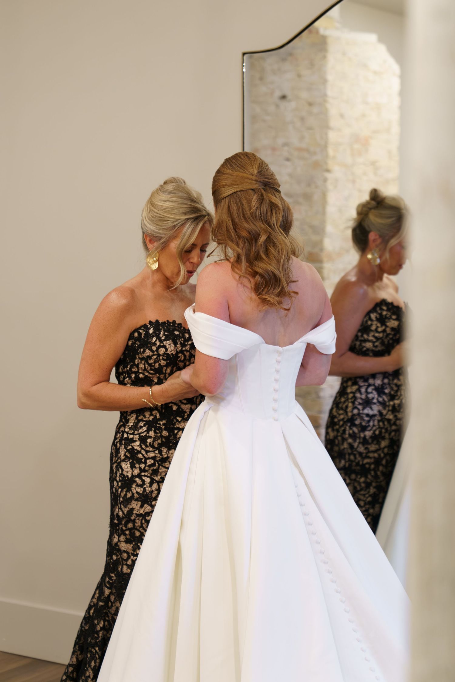 A bride in a white off-shoulder gown gets her buttons fastened by a woman in black lace dress, reflected in a mirror.