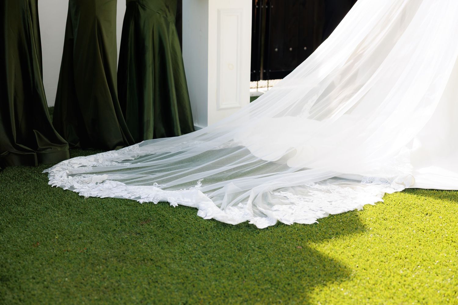 White wedding dress train spread on green grass with dark olive bridesmaid dresses in background.