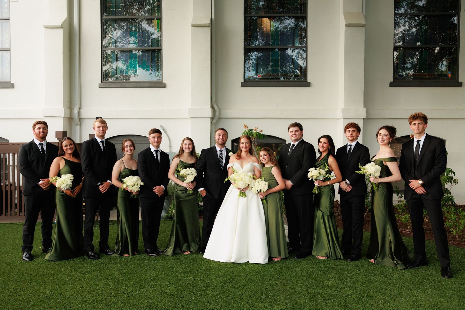 Wedding party in green dresses and black suits posing outdoors in front of a white building with bride in white gown.