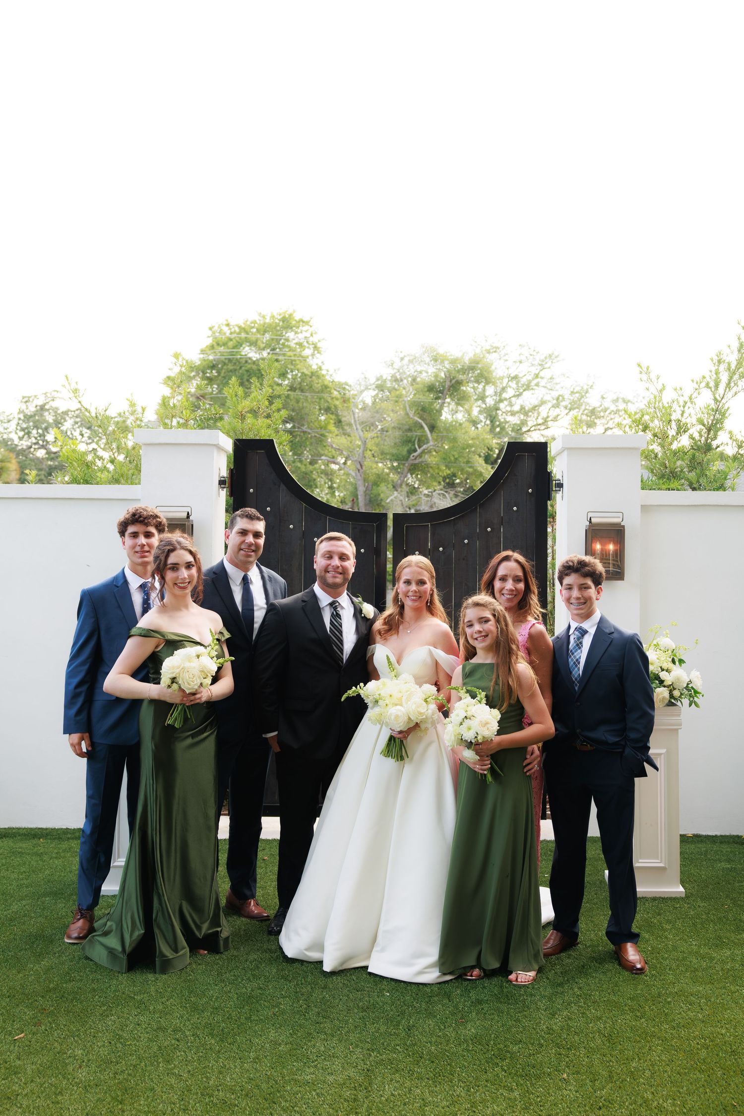 Wedding party poses outdoors in green bridesmaid dresses and navy suits before black gates.
