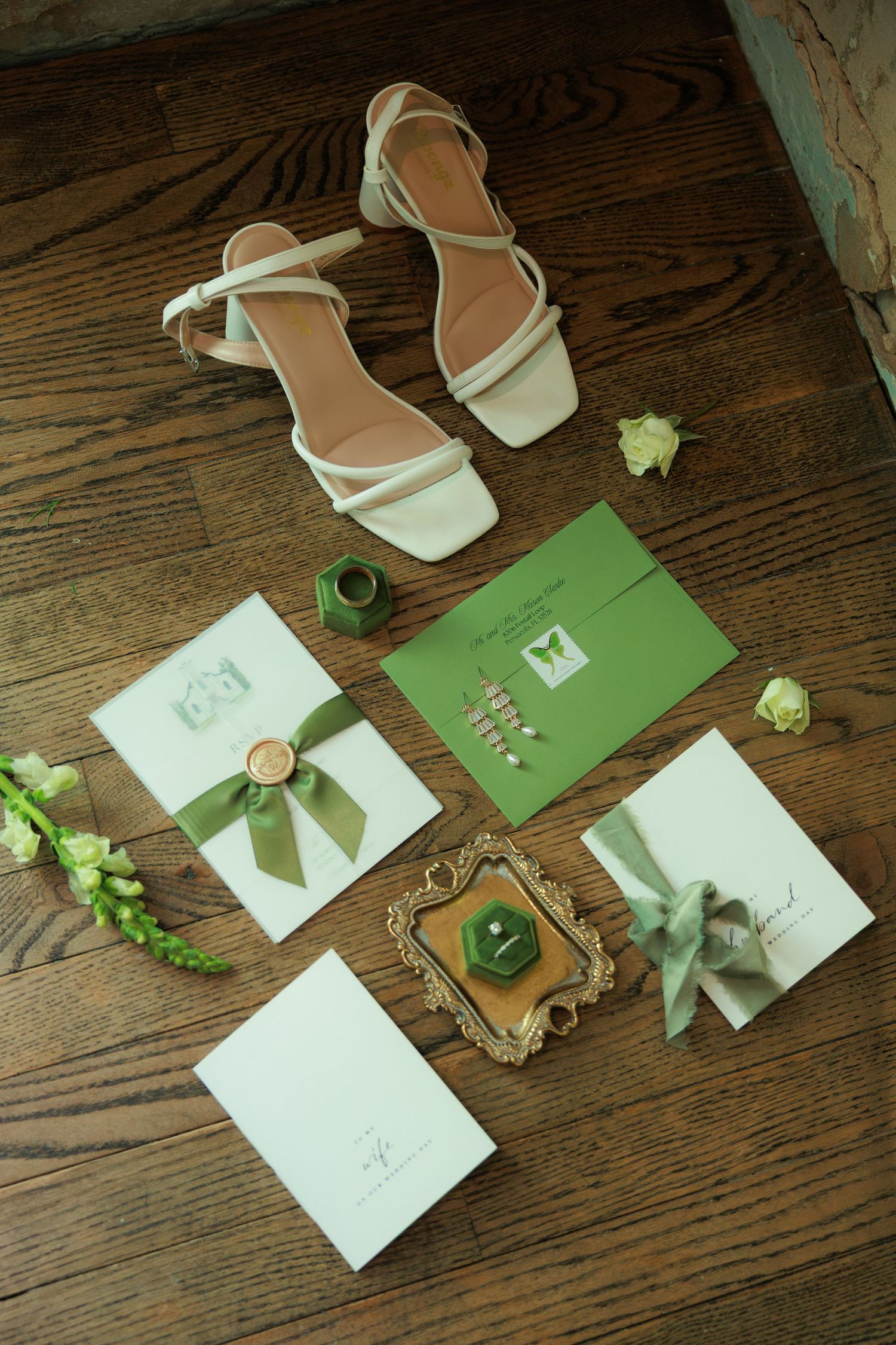 White wedding heels with green ribbon stationery suite, ring box, and floral accents arranged on rustic wood floor.
